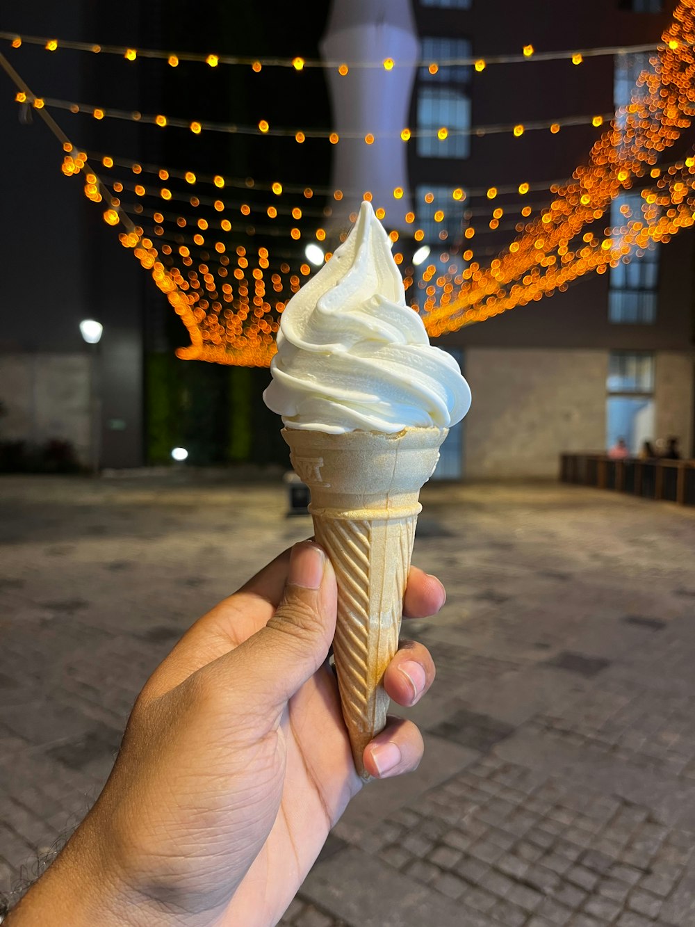 a hand holding an ice cream cone with a string of lights in the background