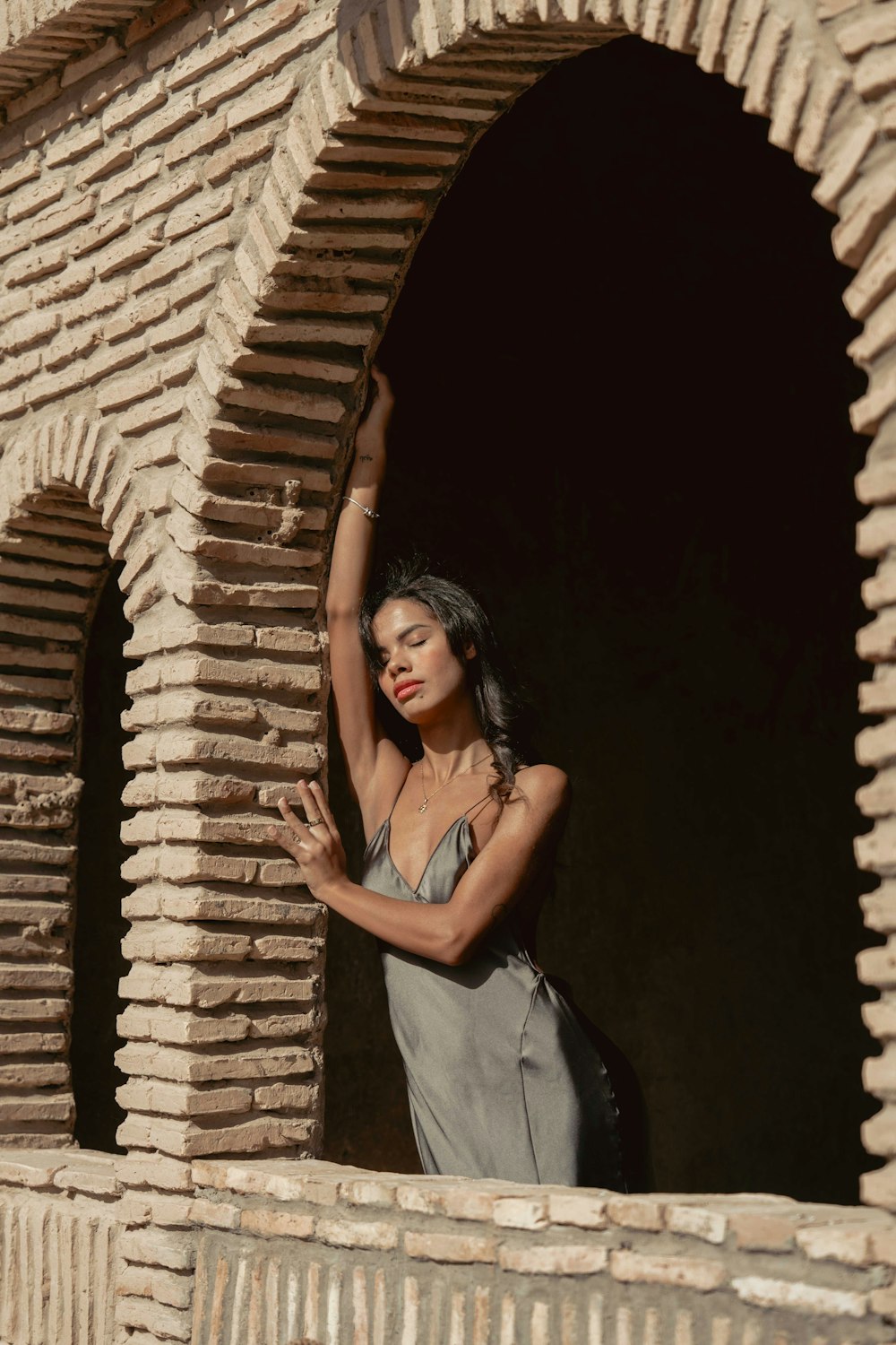 a woman in a gray dress leaning against a brick wall