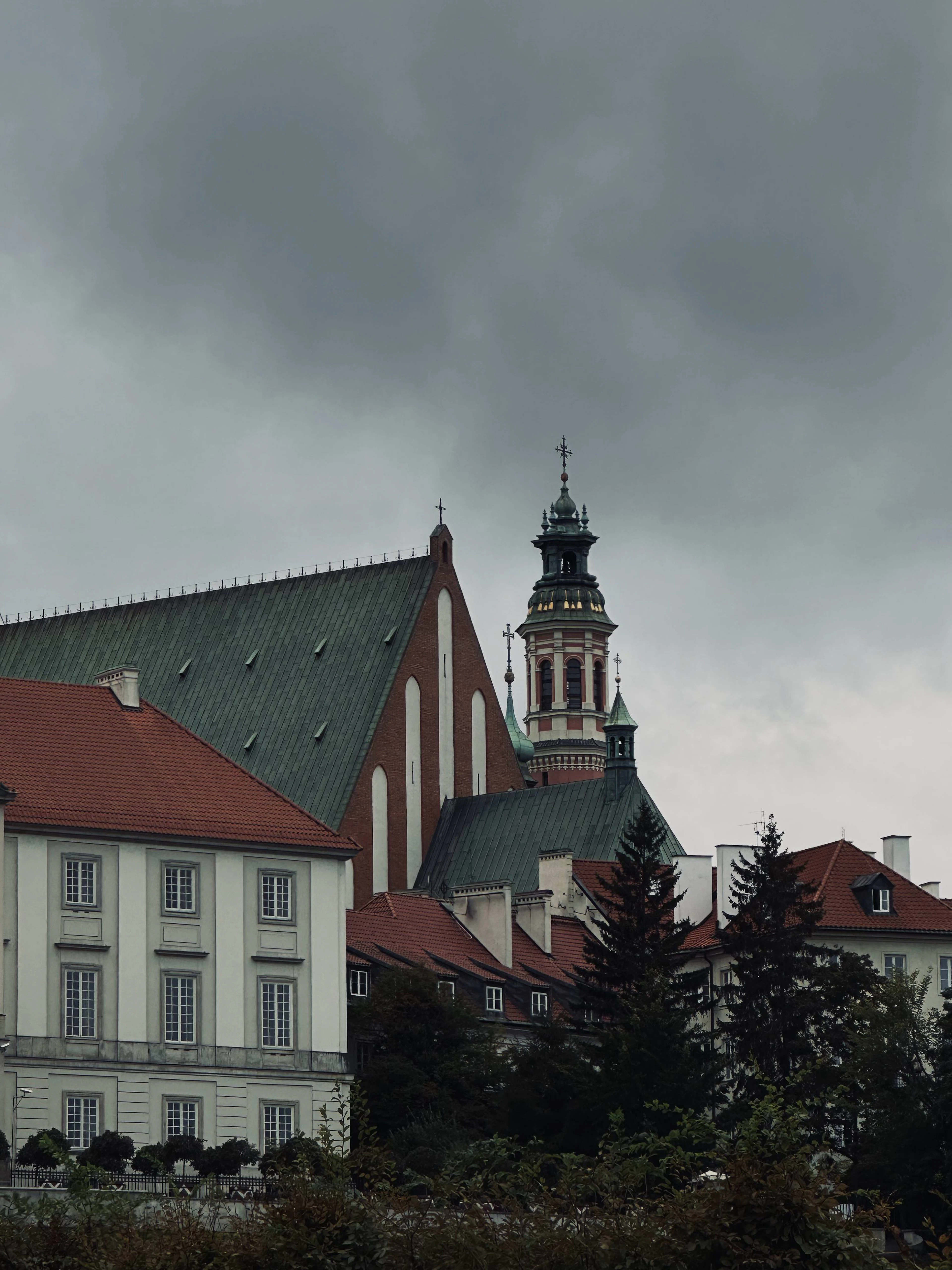 A baroque church tower with a green dome rises above Warsaw's Old Town rooftops under a stormy sky.