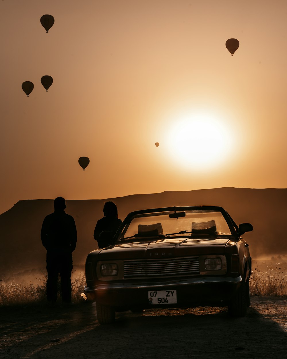 two men standing next to a car with hot air balloons in the sky