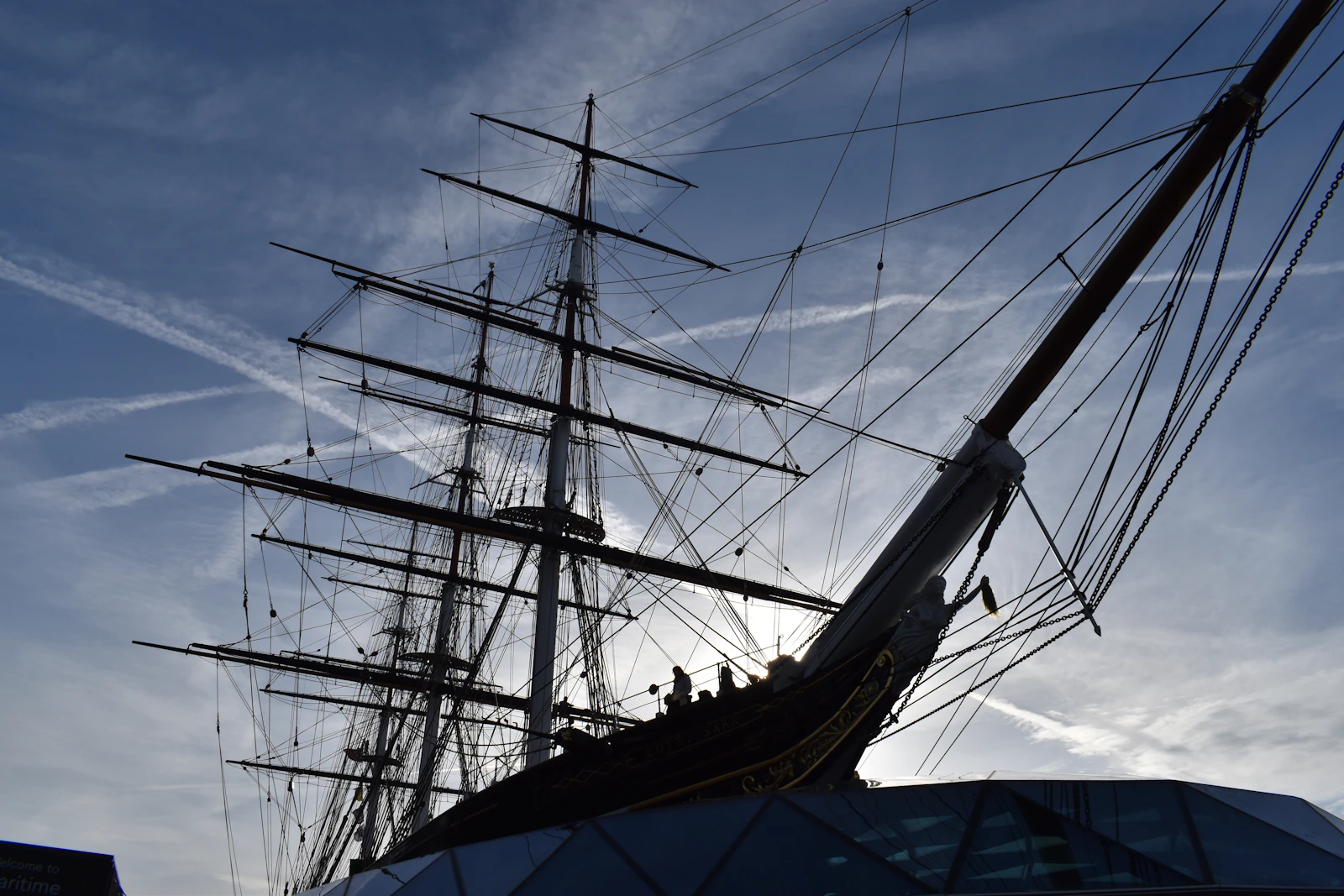 Photograph of Cutty Sark’s dark bow, masts and web of rigging rise against a streaked blue sky in Greenwich.