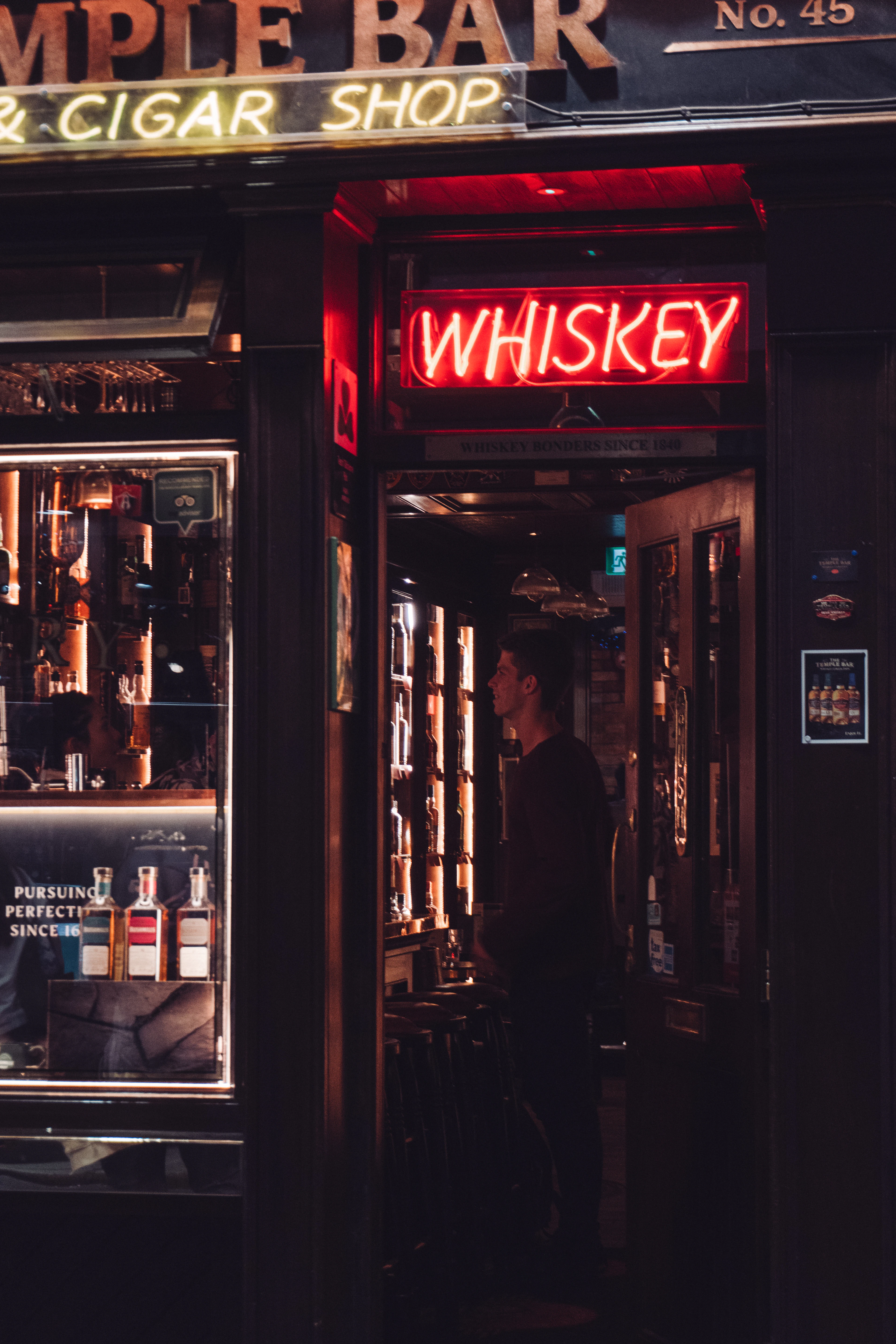 A red neon Whiskey sign glows above a bar doorway at night with a figure inside.
