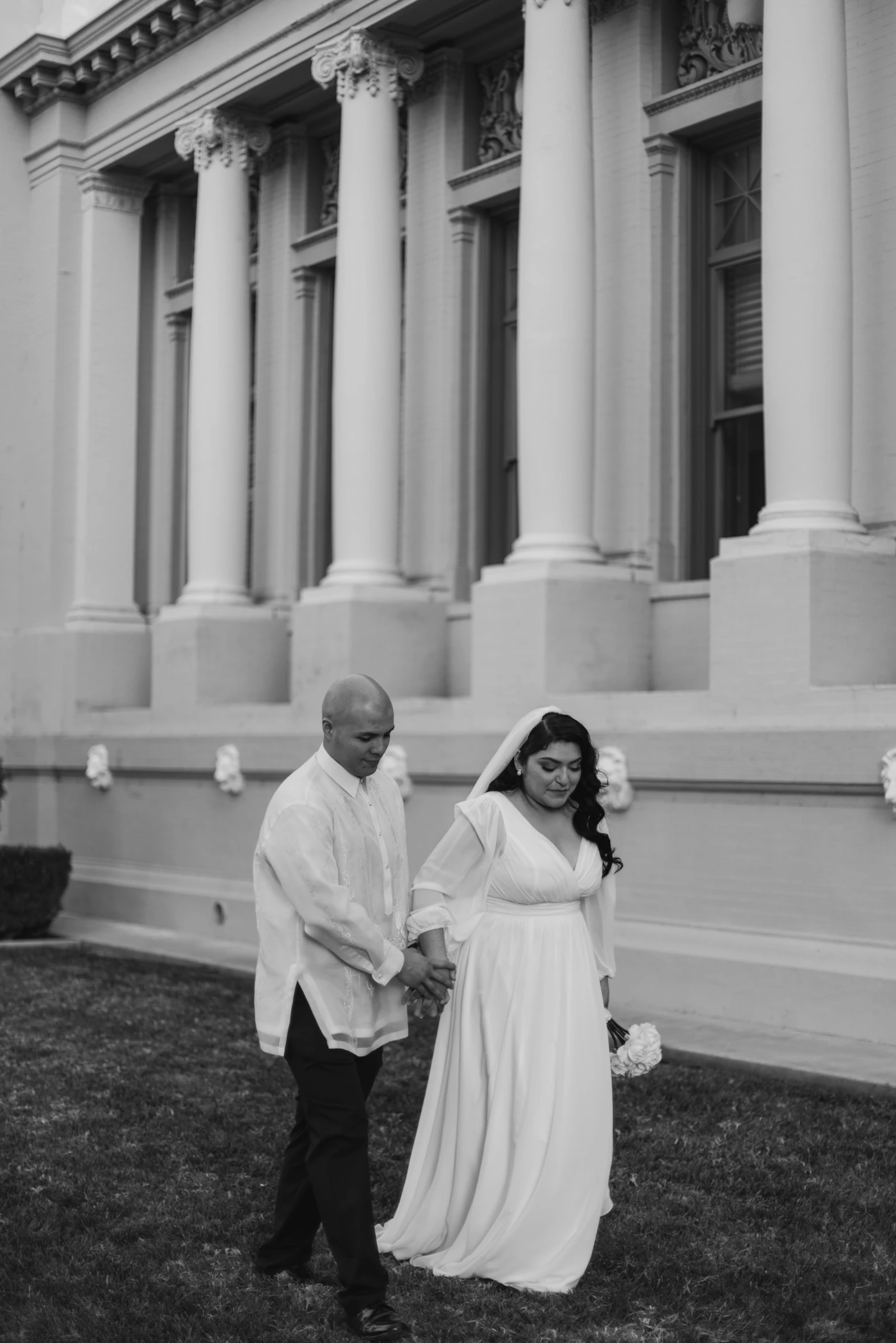 Bride and groom walking hand in hand along wide path in front of grand stone building with soft natural light