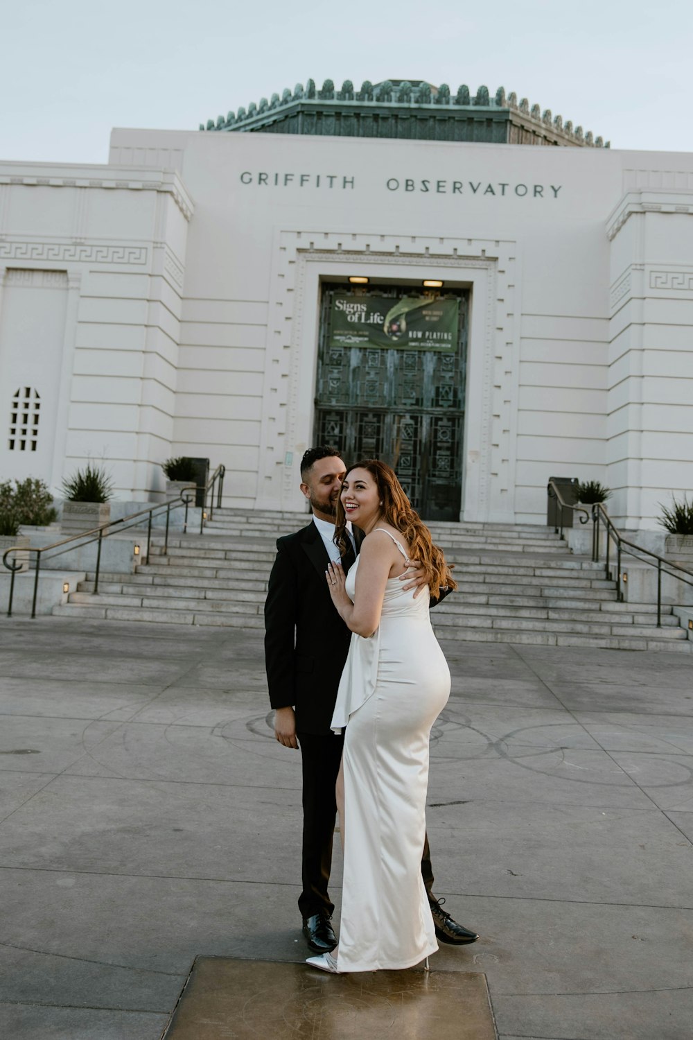 a pregnant woman in a white dress standing next to a man in a black suit