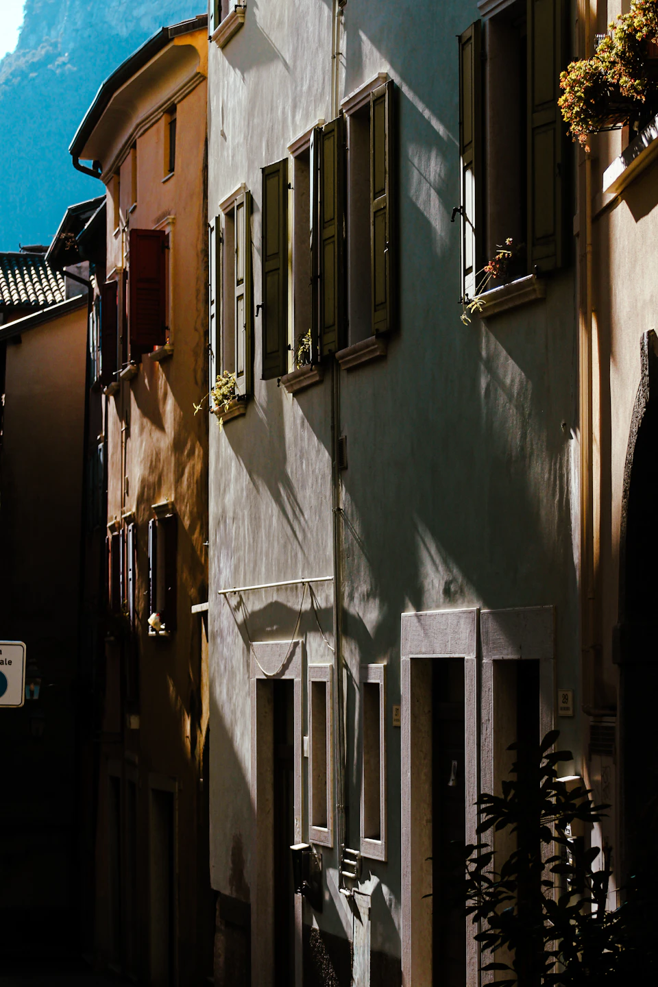 The image depicts a narrow street lined with old, multi-story buildings painted in shades of white and beige. The buildings feature wooden shutters on their windows, adding character to the scene. A few potted plants are visible on the ground level, providing some greenery amidst the urban landscape. The perspective of the photo is taken from a low angle, looking up at the buildings, which enhances their imposing presence against the backdrop of the clear blue sky with scattered clouds.

The image does not contain any discernible text or countable objects, and there are no people visible in the frame.