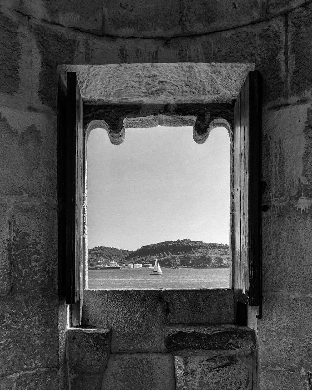 a window in a stone wall with a sailboat in the distance