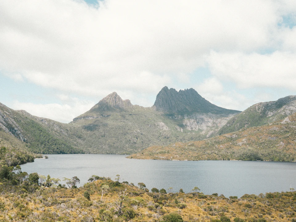 The image depicts a serene mountain landscape with two prominent mountains, one on each side of a large lake. The mountains are covered in lush green vegetation and have a rugged appearance. The sky above is filled with clouds, casting a soft light over the scene. The perspective of the photo is from a distance, providing a comprehensive view of the natural beauty of the landscape.
