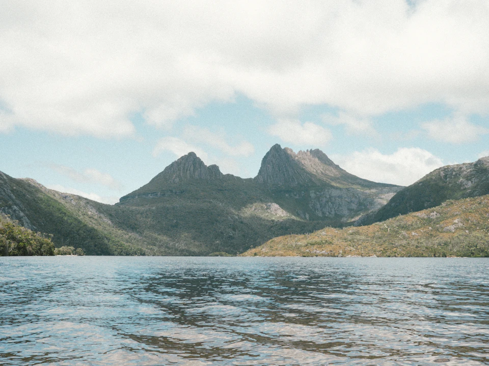 The image depicts a serene mountain landscape with two large, dark gray mountains on either side of a light blue lake. The mountains are covered in lush green vegetation and have a rugged appearance. The sky above is filled with clouds, creating a dramatic backdrop for the scene. The perspective of the photo suggests it was taken from the water, looking towards the mountains.