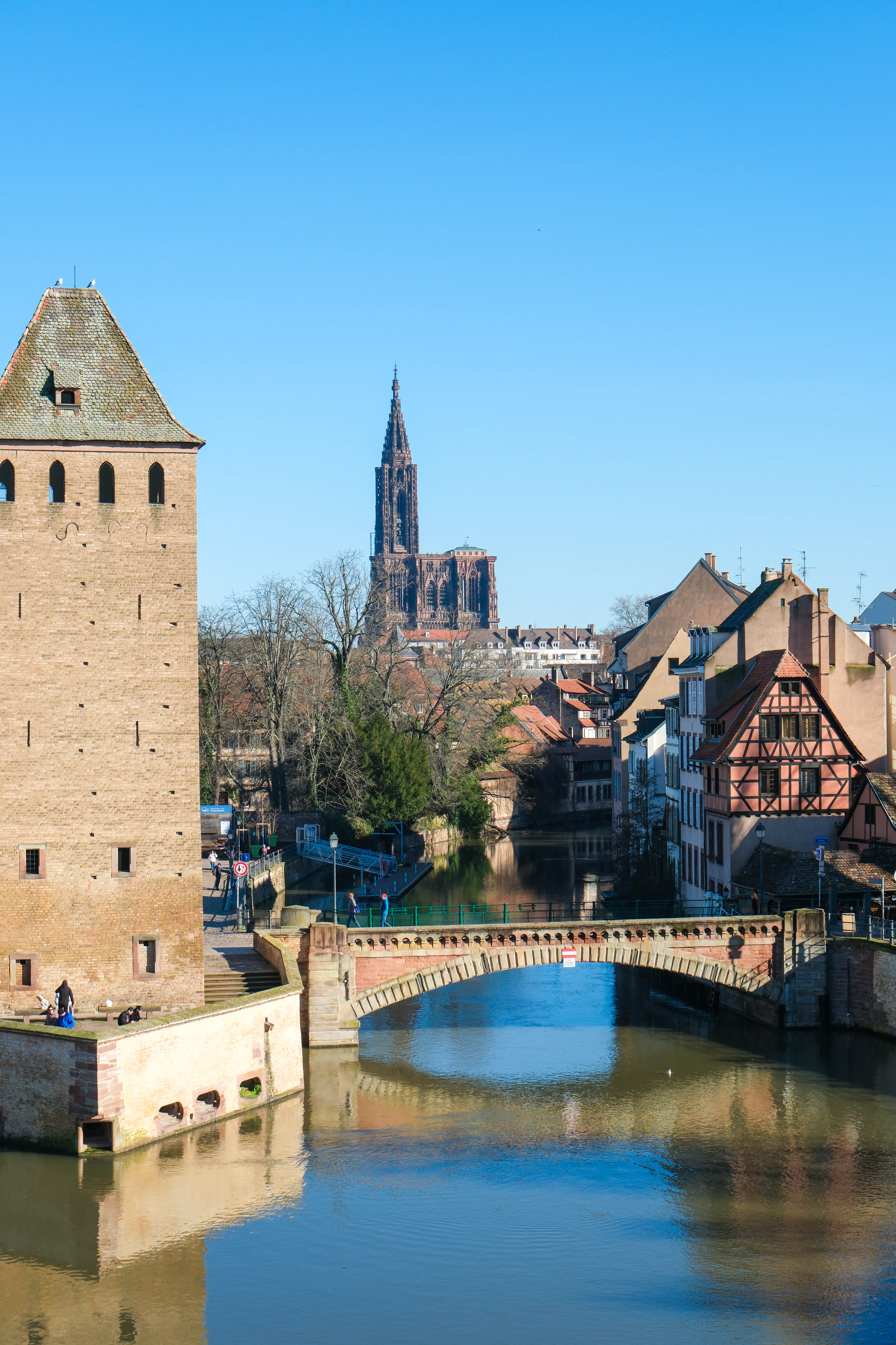 One of the medieval watchtowers of the Ponts Couverts standing over the River Ill in Strasbourg, half-timbered houses on the far bank and the cathedral spire rising behind under a clear blue sky.