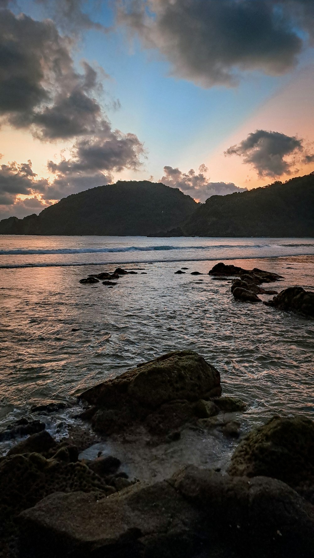 the sun is setting over the ocean with rocks in the foreground