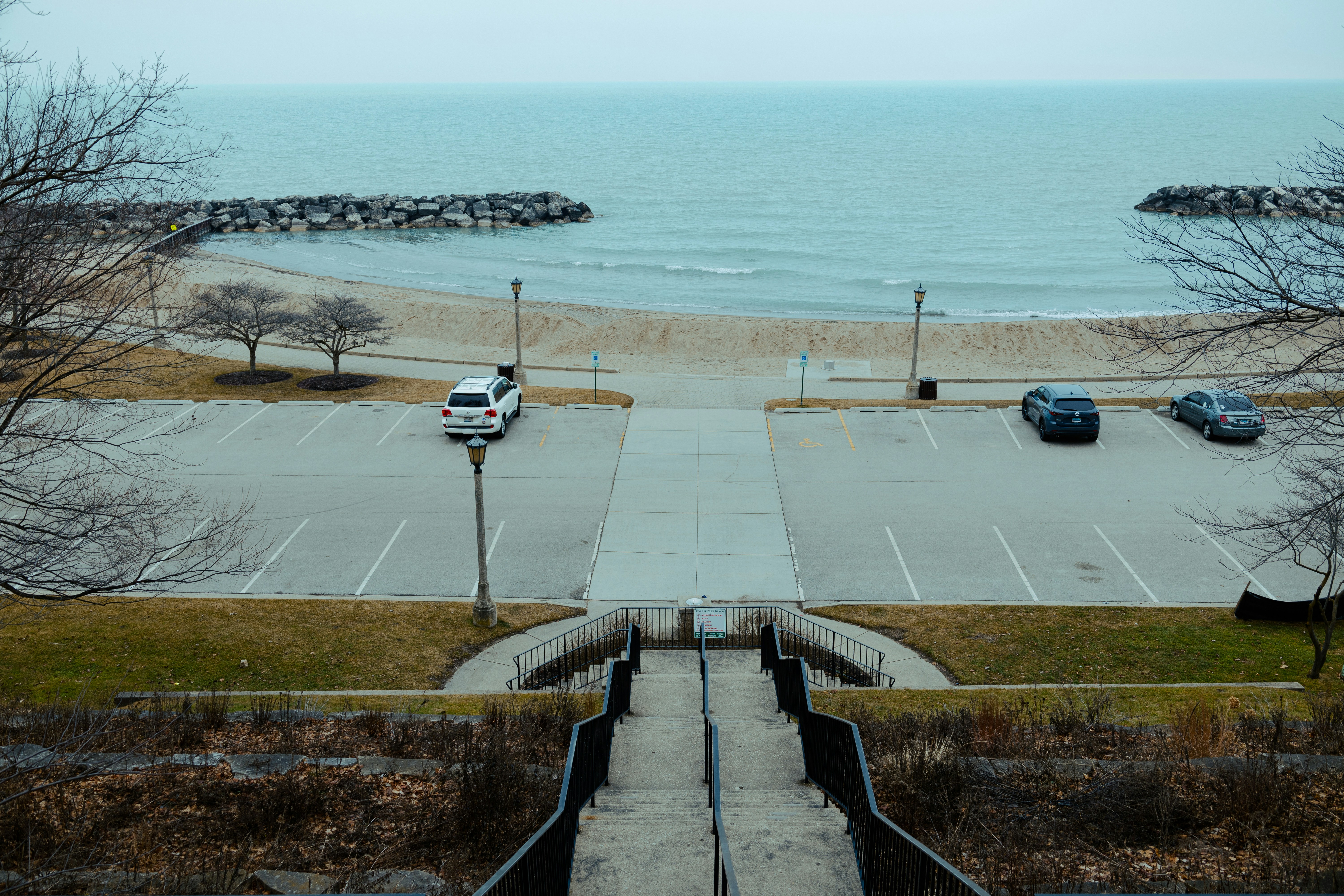 Photograph of a symmetrical staircase descending toward a calm sea, framed by a vacant parking lot and bare trees.