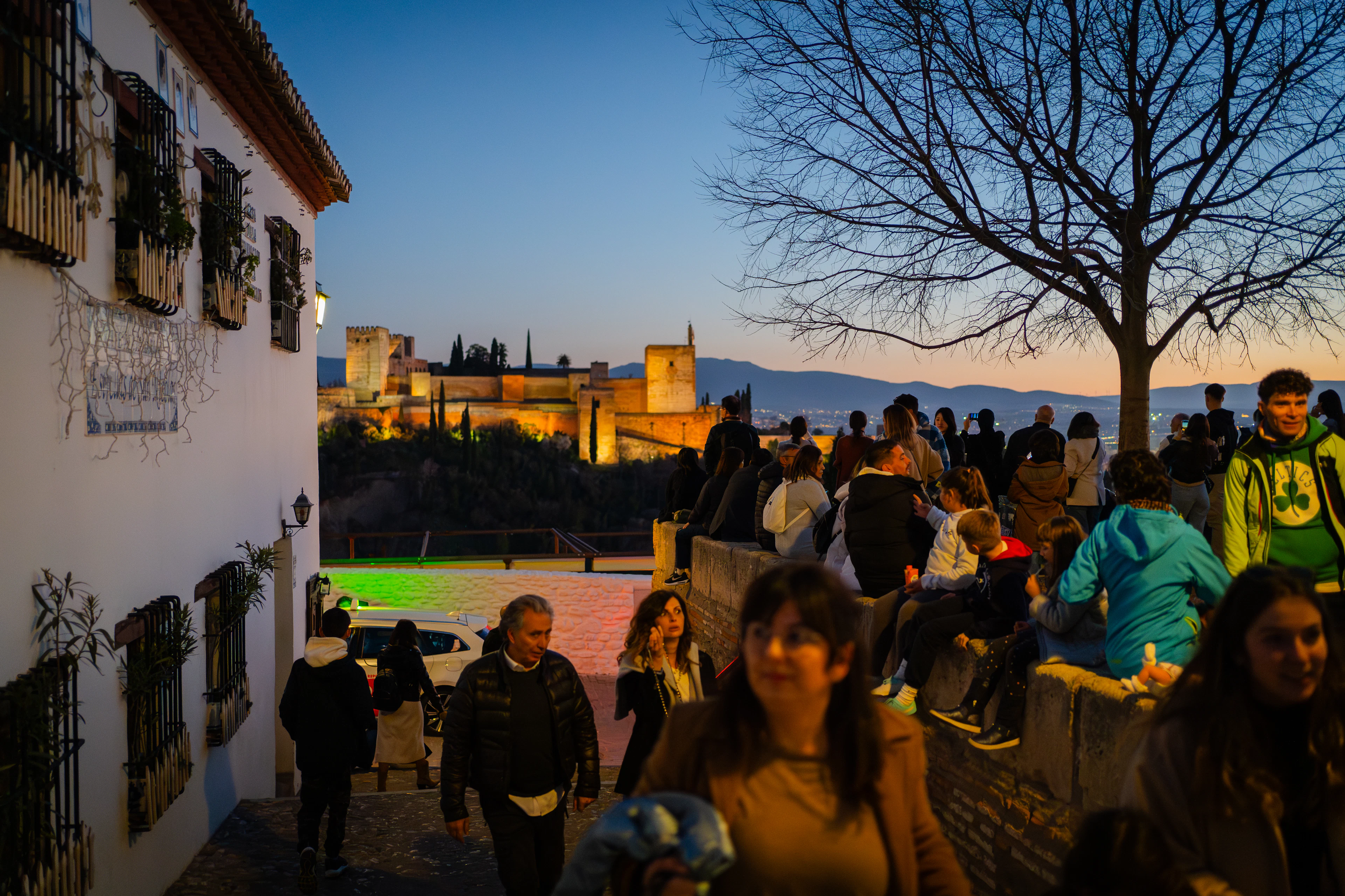 Crowds gather at a Granada viewpoint at dusk as the illuminated Alhambra glows across the valley.