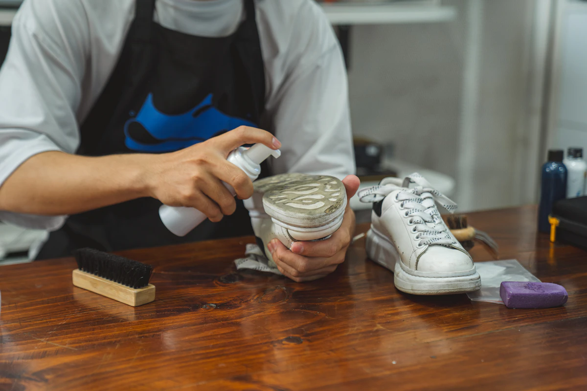 Un artisan en tablier noir vaporise un produit sur la semelle d'une basket blanche, au-dessus d'un comptoir en bois où attendent l'autre chaussure, une brosse et un pain de nettoyage