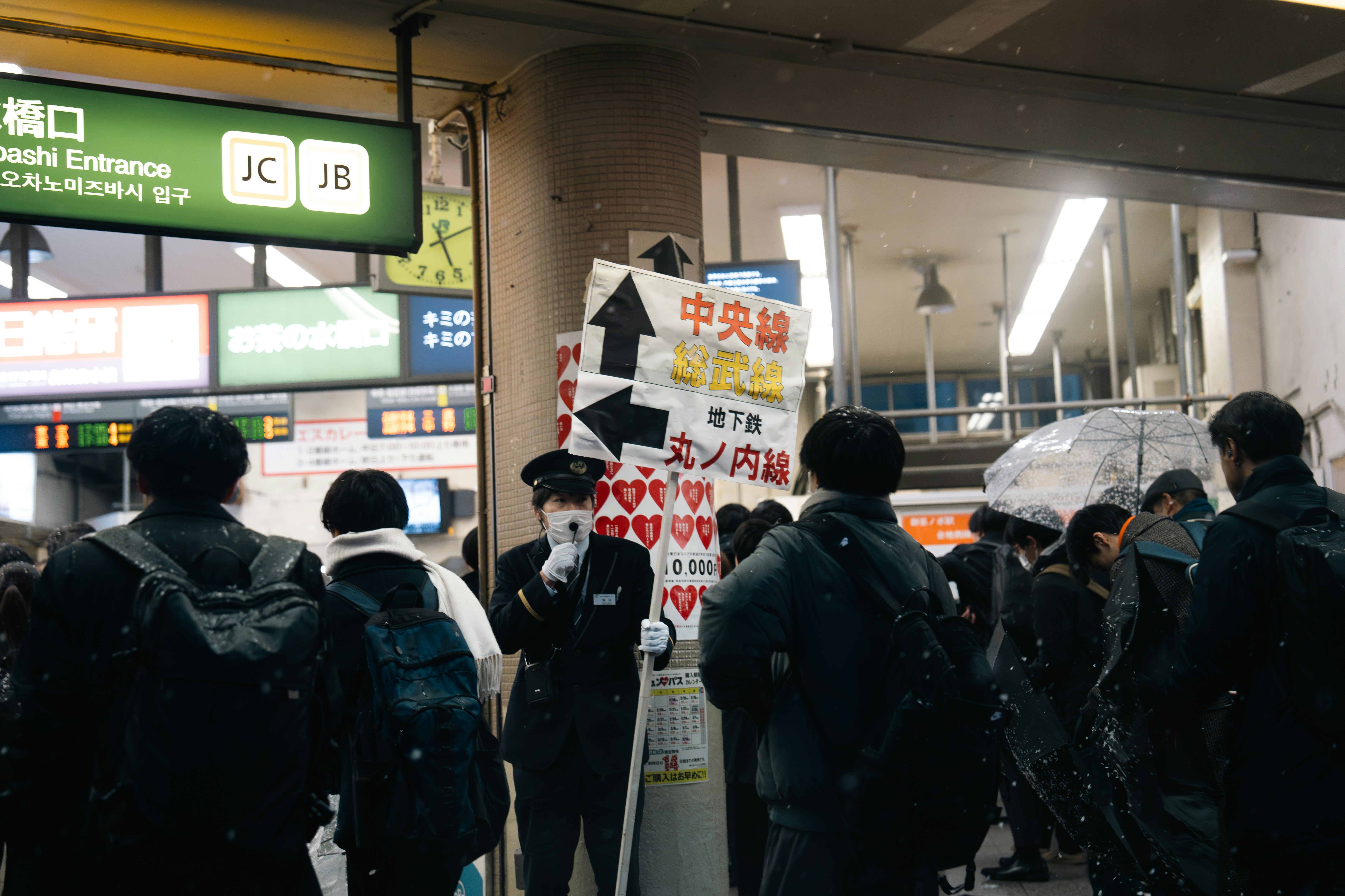 a group of people standing next to each other holding umbrellas, In the midst of a heavy snowfall at Ochanomizu Station, Tokyo, a station attendant diligently informs passengers of train schedule disruptions while guiding them to alternative transportation options.