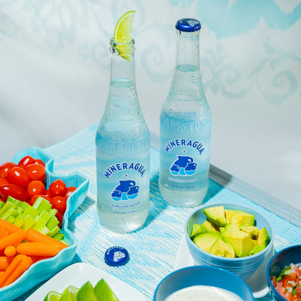 two bottles of mineral water sitting next to bowls of fruit and vegetables