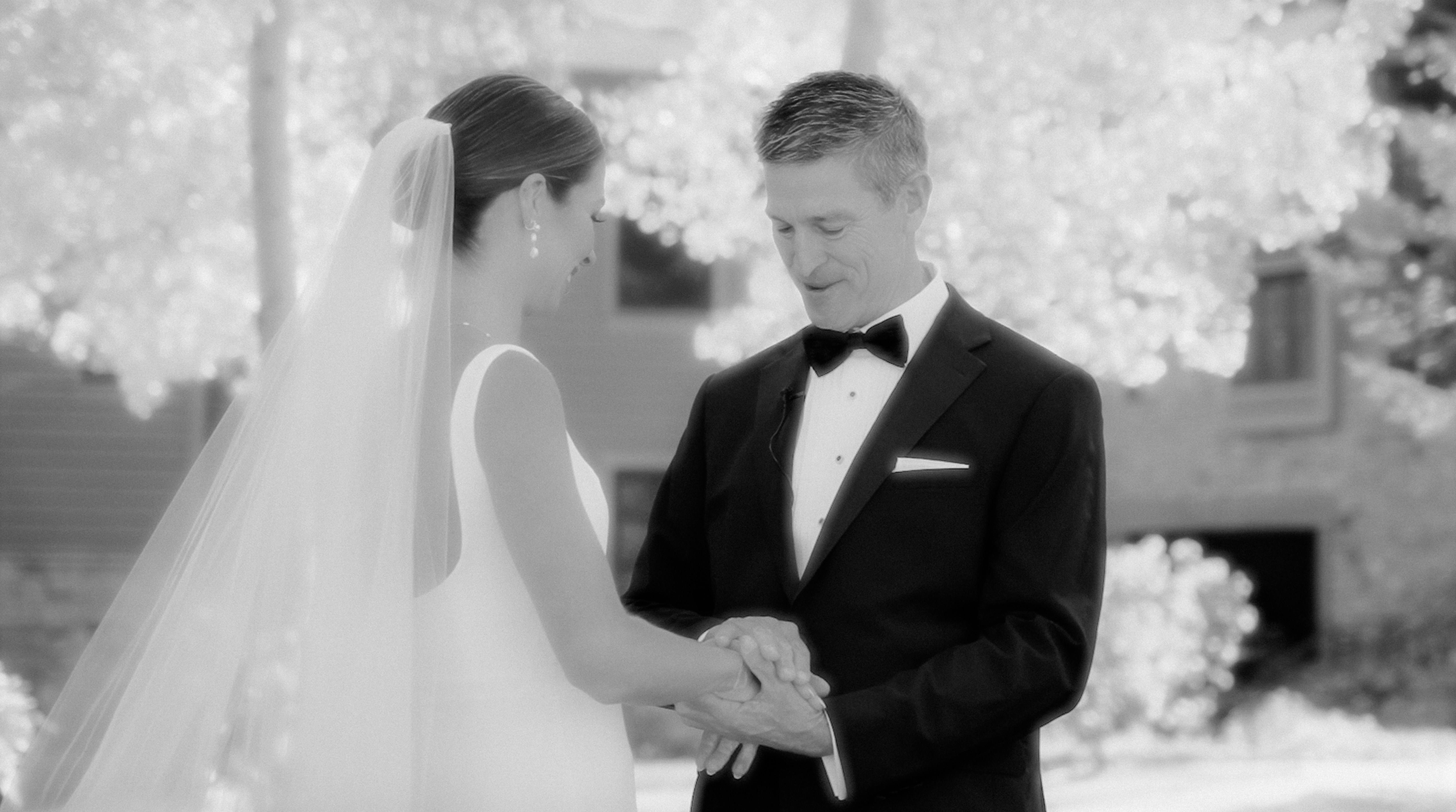 Bride and groom holding hands at ceremony, warm natural light, blurred floral background, intimate close-up