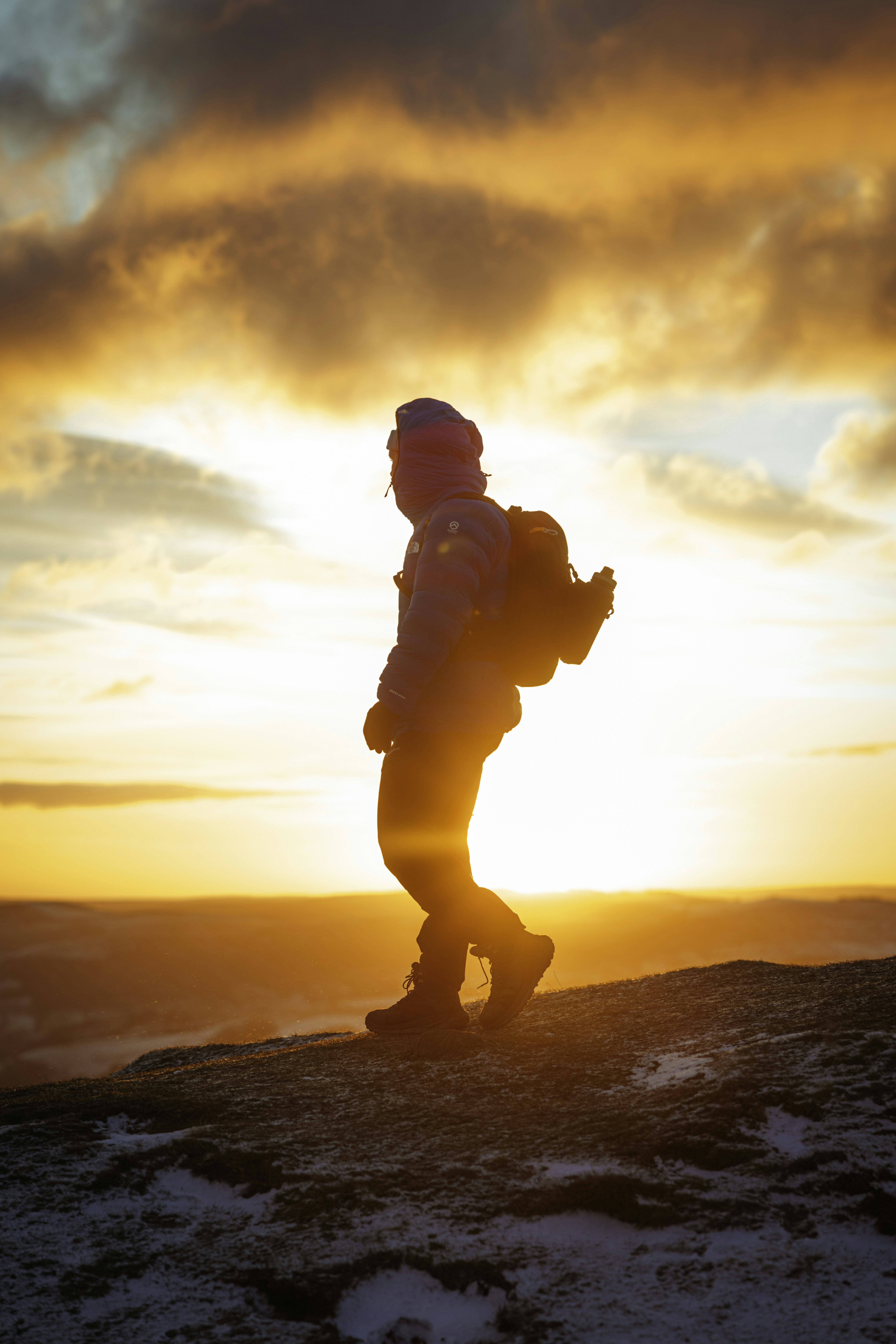 Hiker with backpack on a mountain trail at sunset, dark silhouette, deep orange sky, dramatic landscape