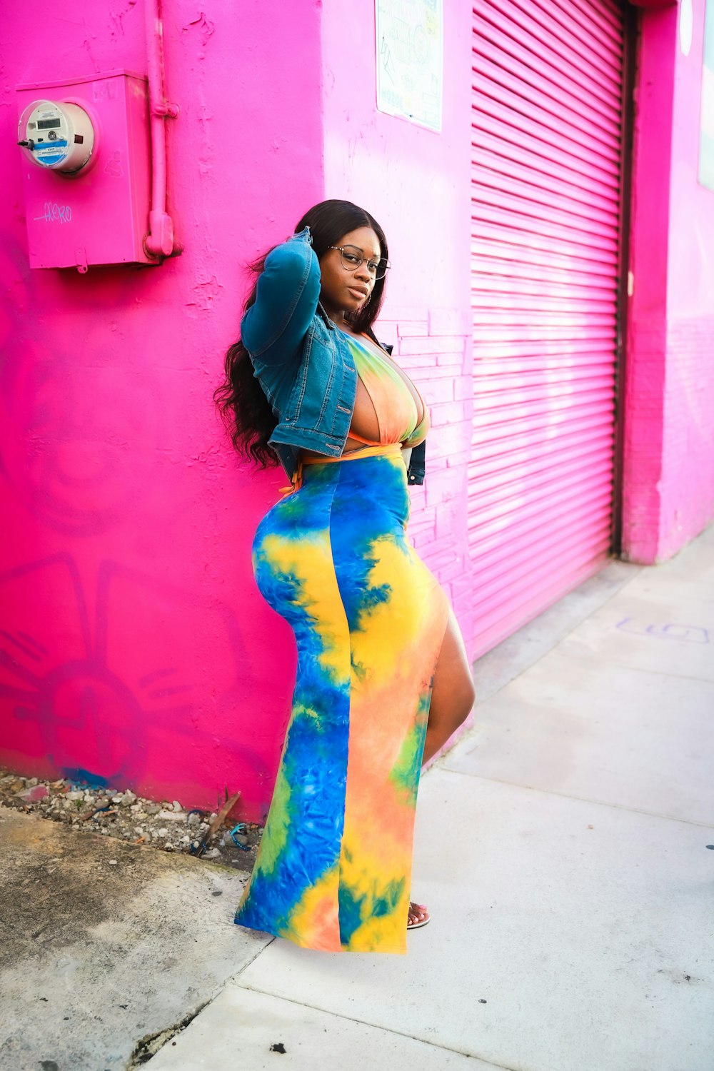 a woman leaning against a pink wall in a tie dye outfit