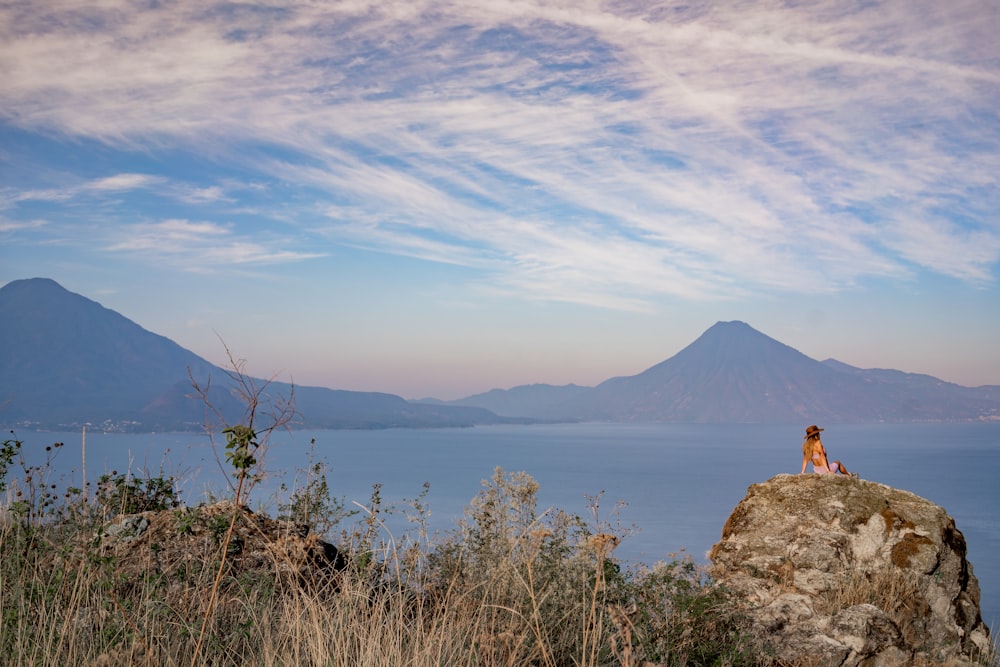 a woman sitting on top of a rock next to a body of water