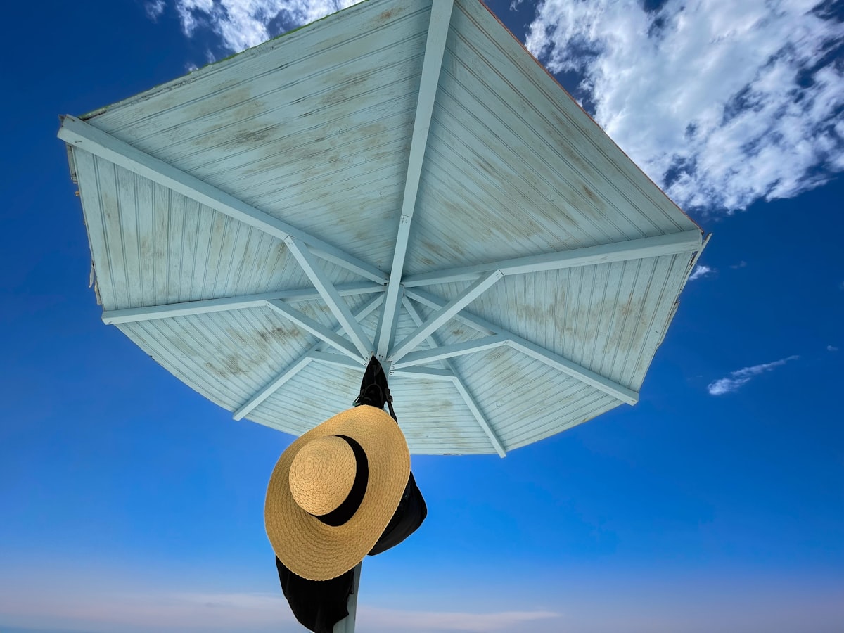 A straw hat hanging under a wooden beach umbrella against a blue sky