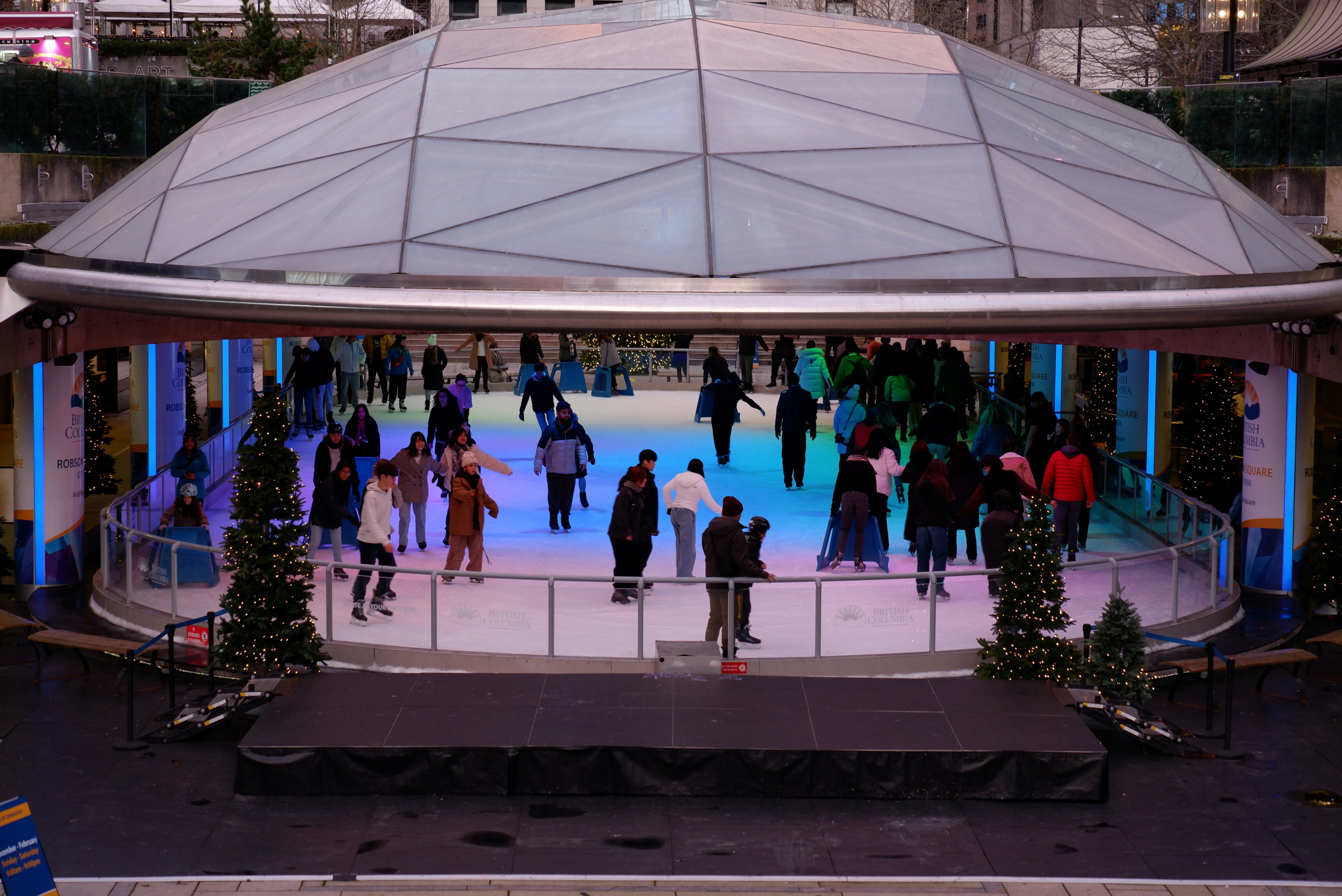 A photo of the Robson Square Ice Rink in Vancouver, Canada. The outdoor ice rink is surrounded by Christmas trees and city lights, with people skating on the ice. (Photo by Itsuka Iwaki on Unsplash)