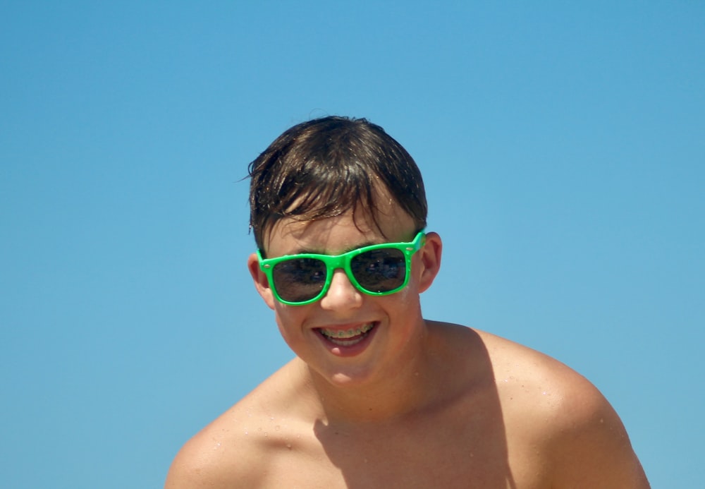 a shirtless boy wearing green sunglasses on a beach