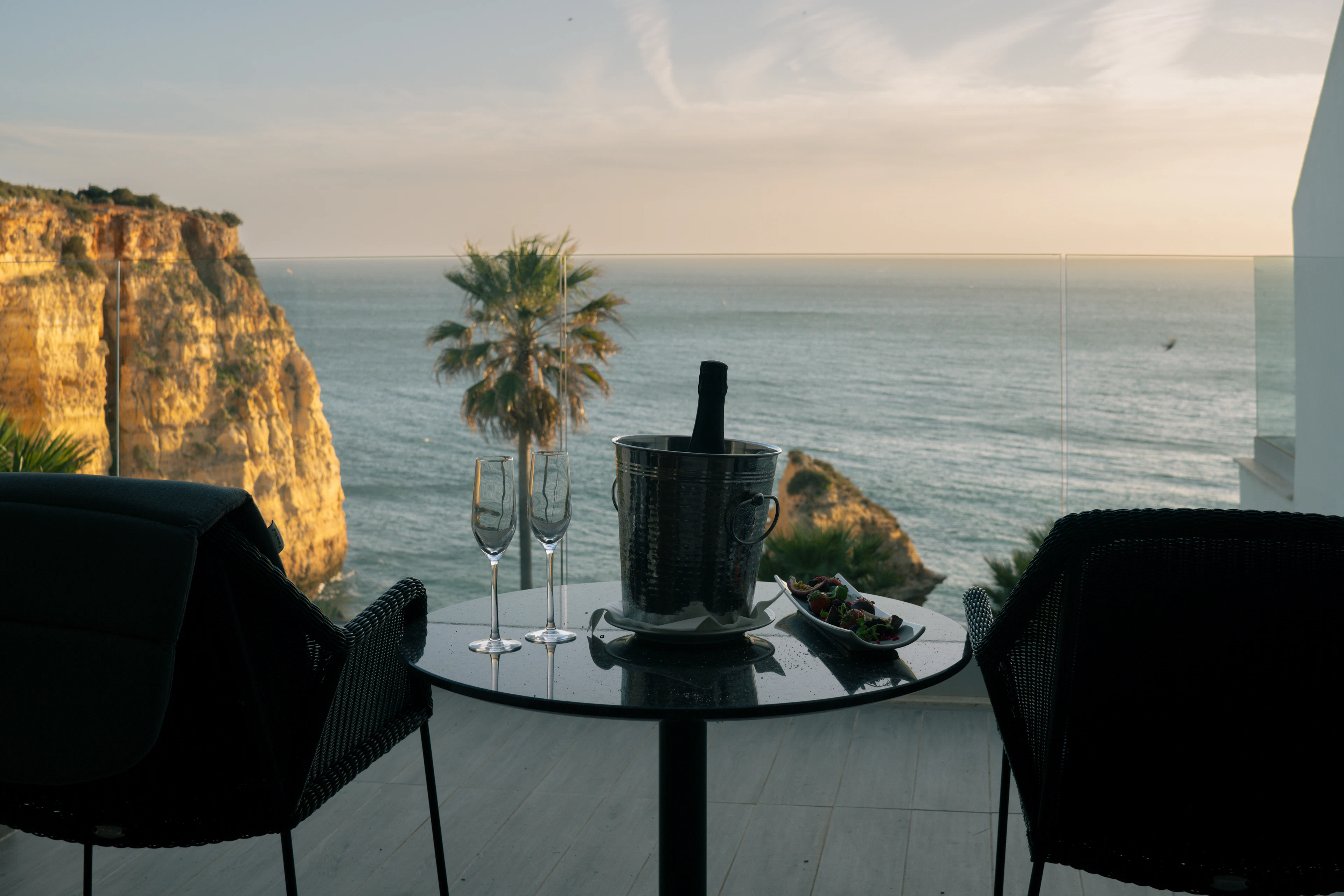 Champagne bucket, two glasses and olives on a terrace table overlooking the Algarve coast at sunset.