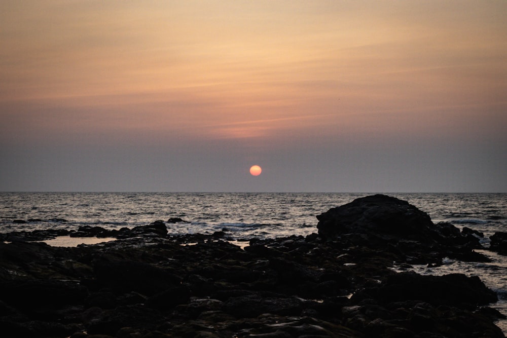 the sun is setting over the ocean with rocks