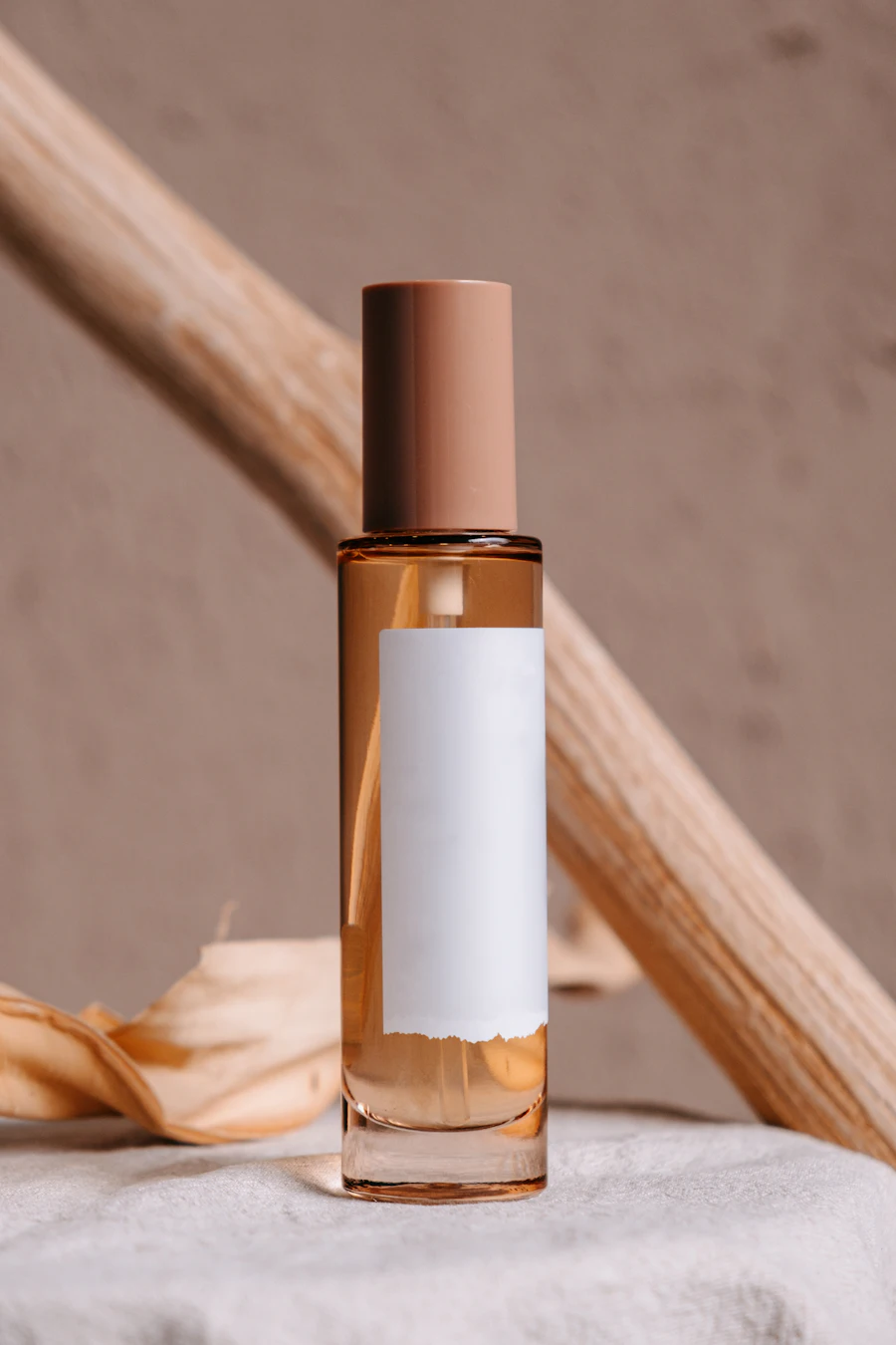 A tall fragrance bottle with a blank label leaning against a branch on a beige backdrop