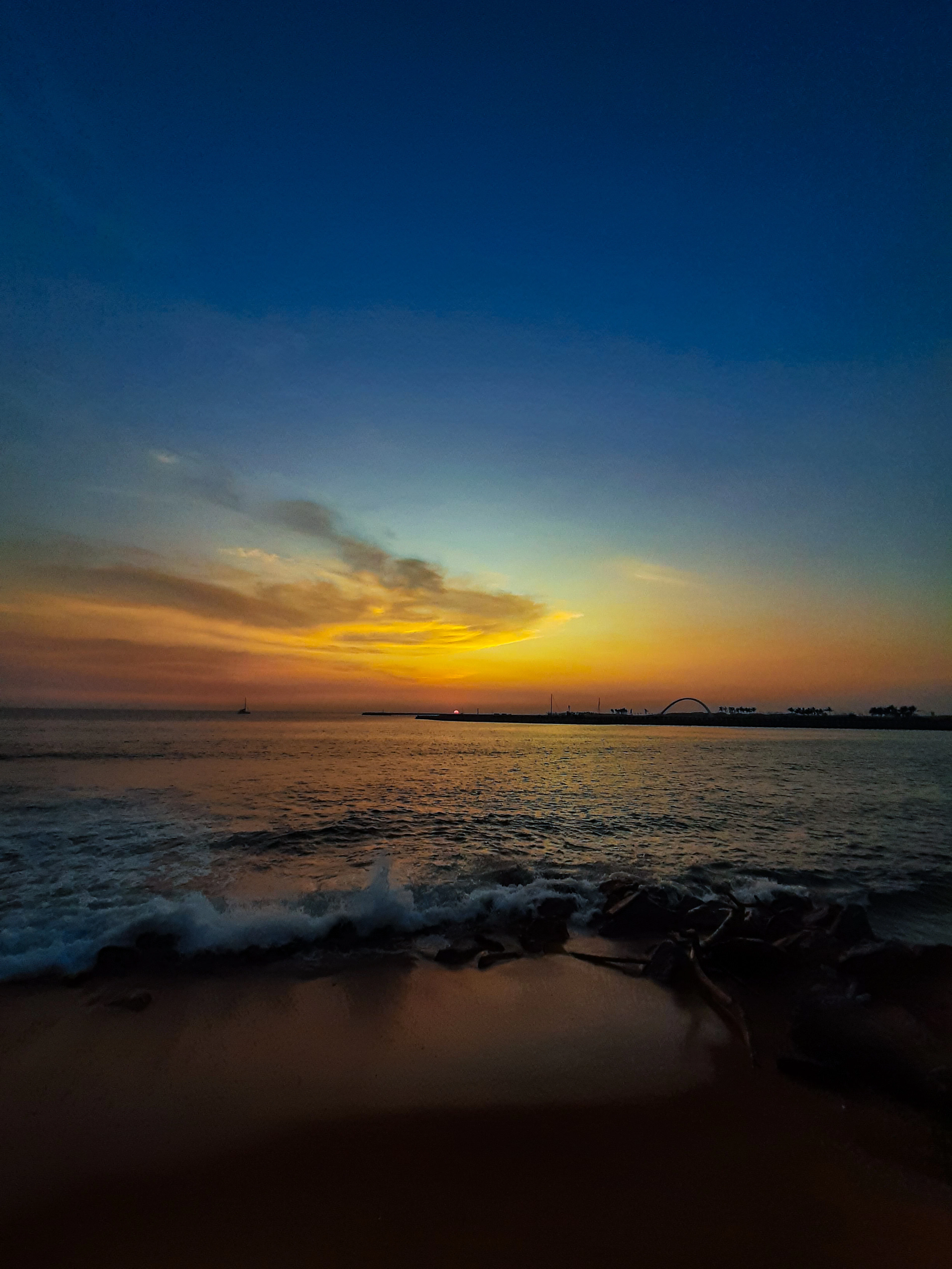 Waves breaking on a beach at dusk under a blue-to-orange sky, a distant bridge silhouette on the horizon.