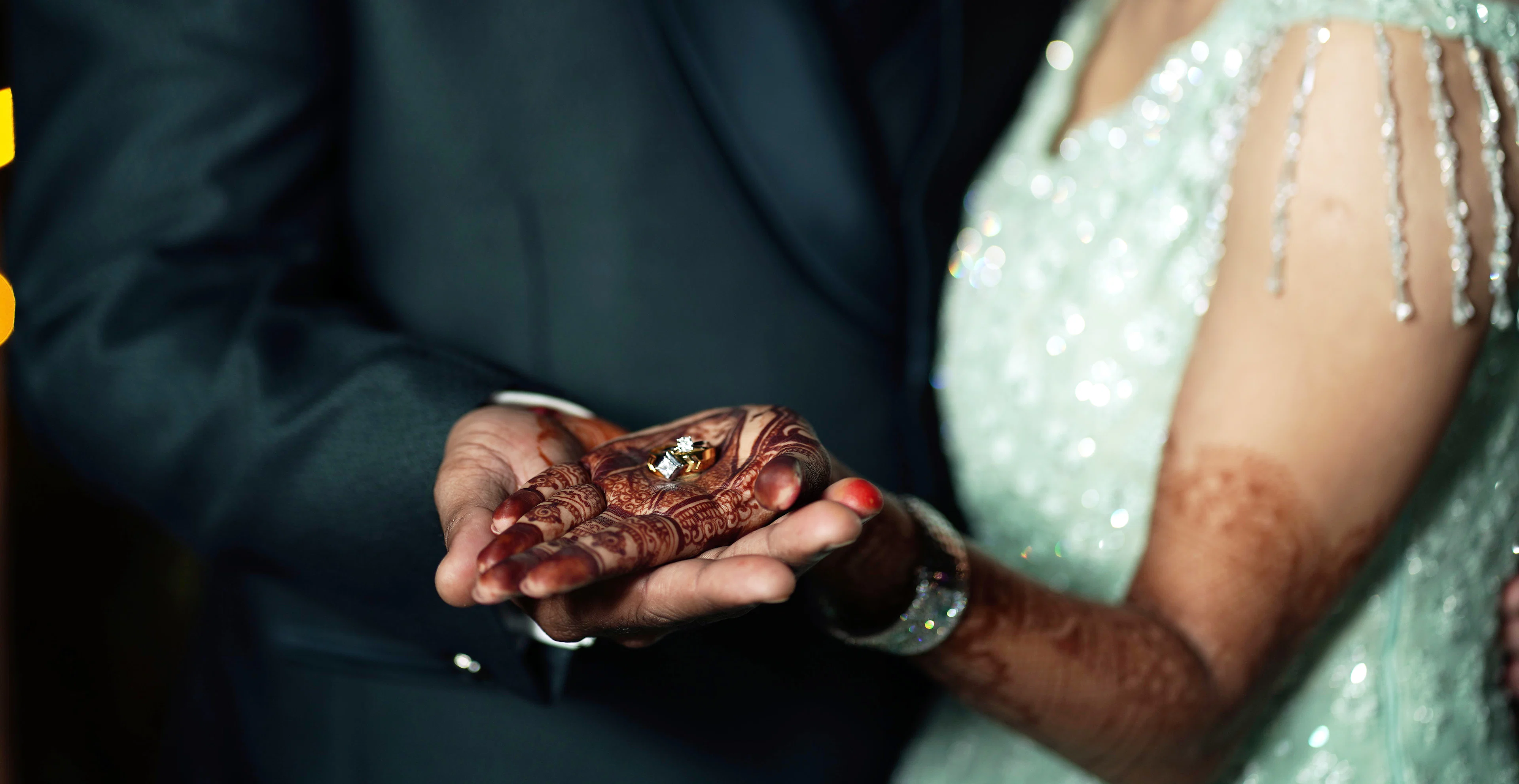 Man in a dark suit holding a woman's henna-patterned hands against her sequinned mint outfit