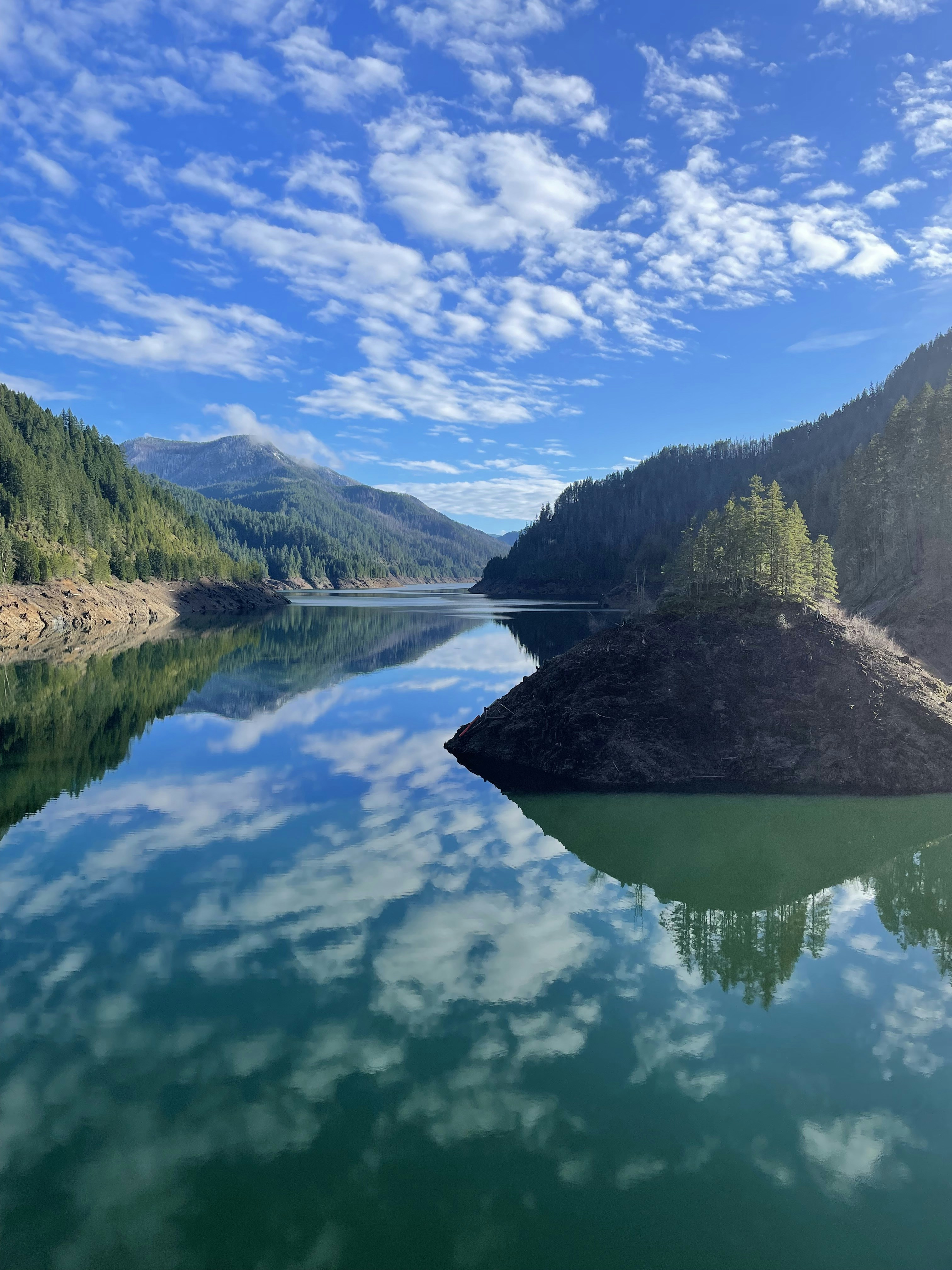 Lac de munte calm reflectând cerul albastru și pădurile verzi, dimineață liniștită fără vânt