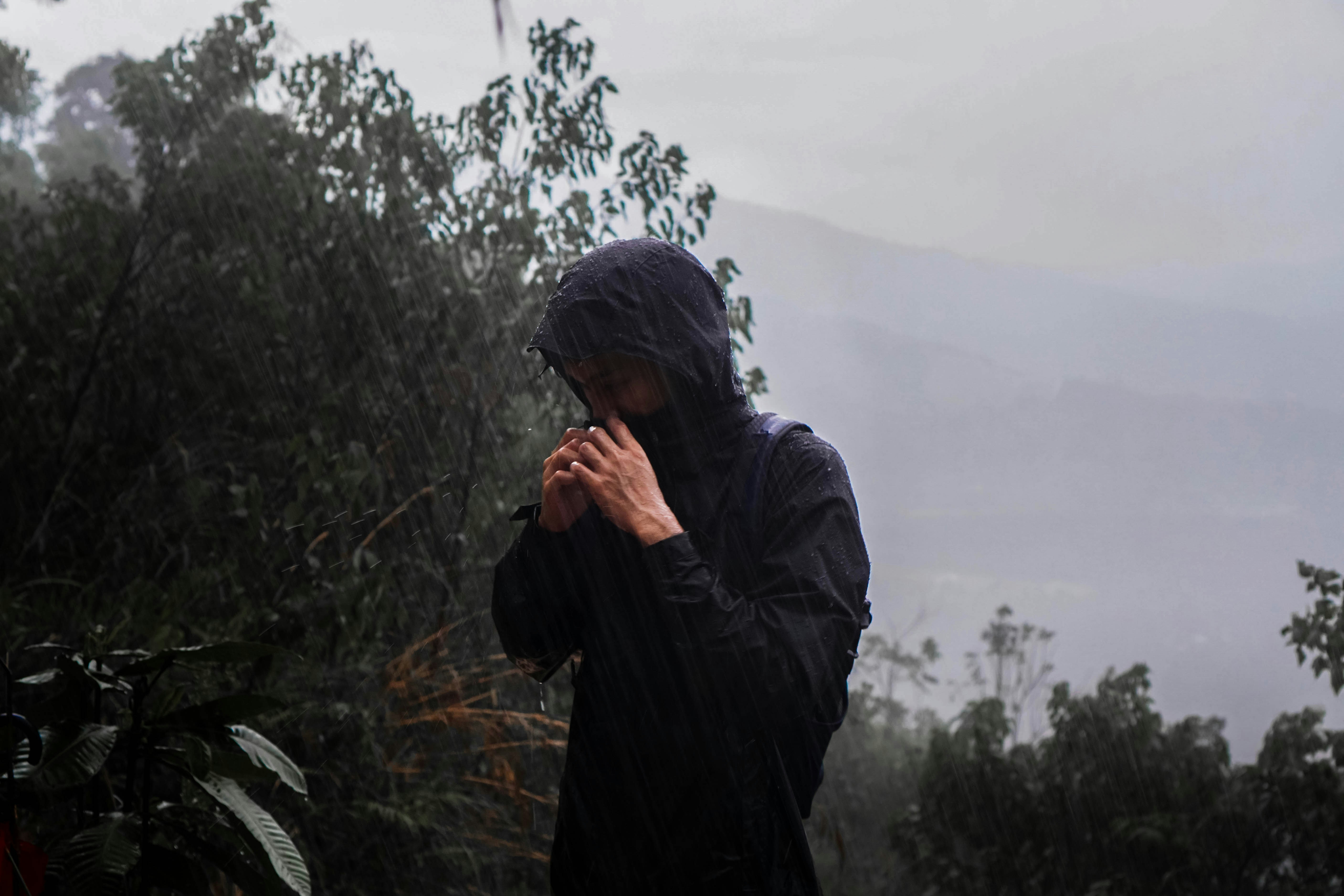 Hiker in a black hooded rain jacket and backpack straps pulling the hood tight against heavy rain on a misty, forested hillside