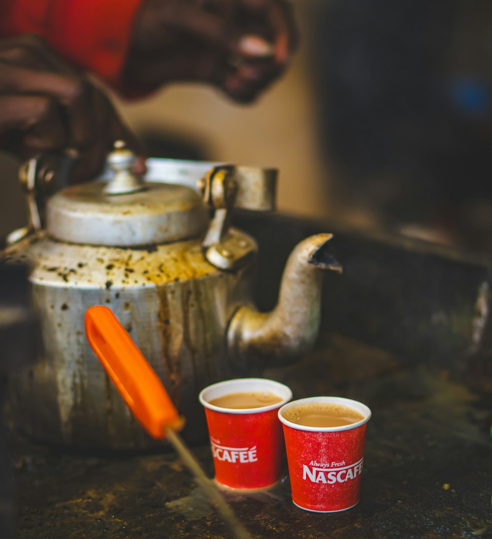 two cups of coffee sit next to a tea kettle