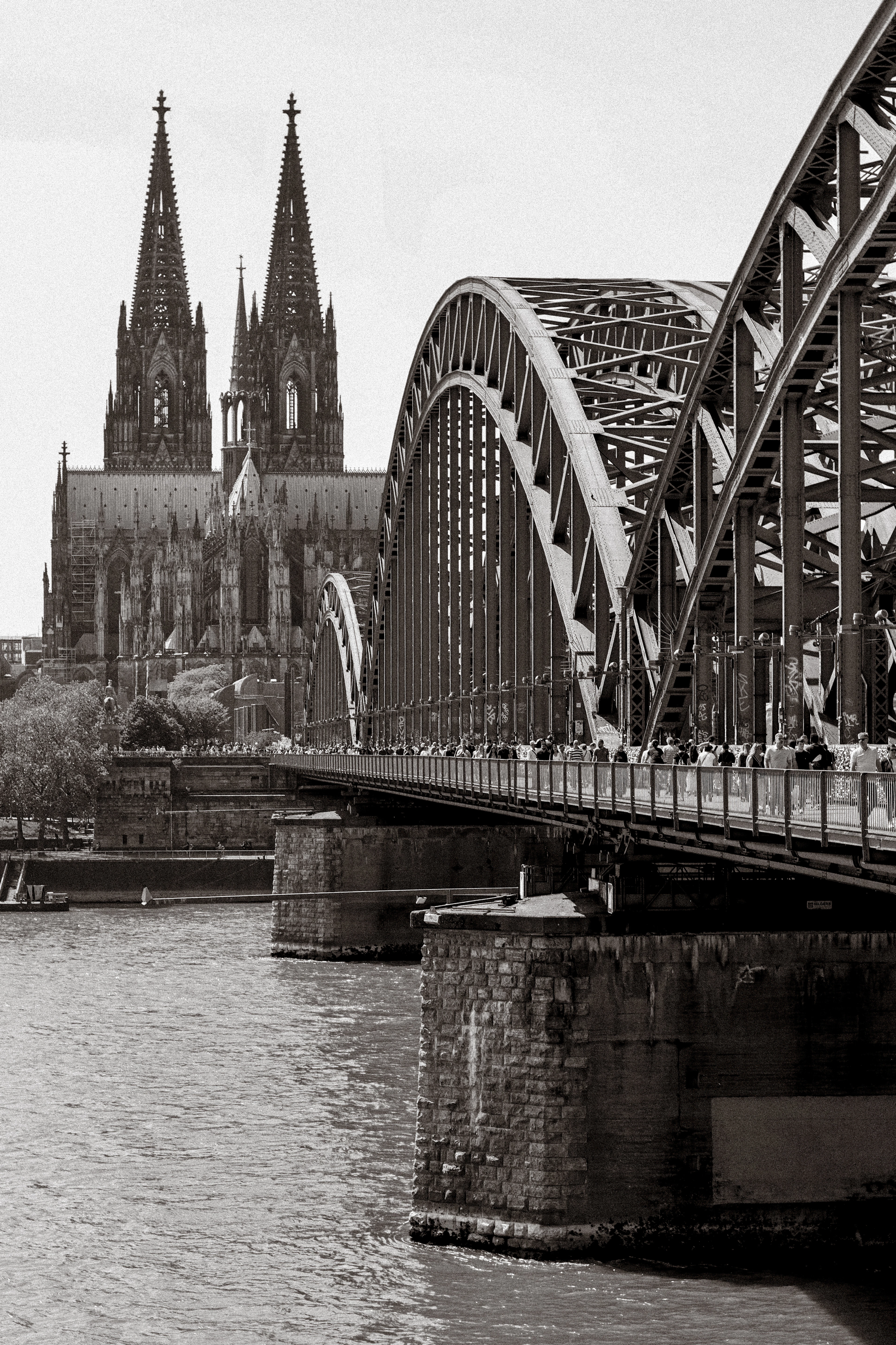Black-and-white view of the Hohenzollern Bridge's steel arch with Cologne Cathedral's twin spires behind it.