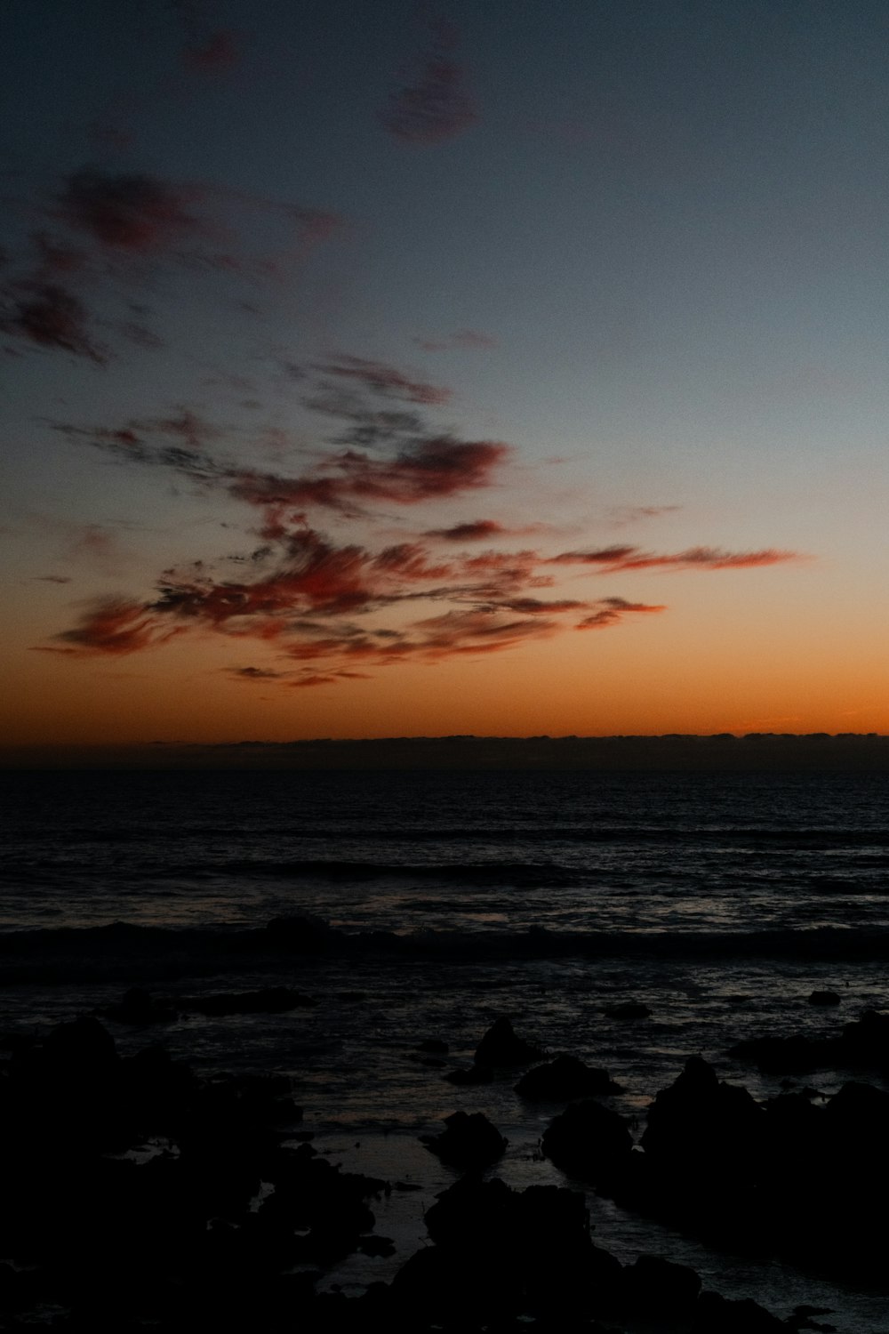 the sun is setting over the ocean with rocks in the foreground