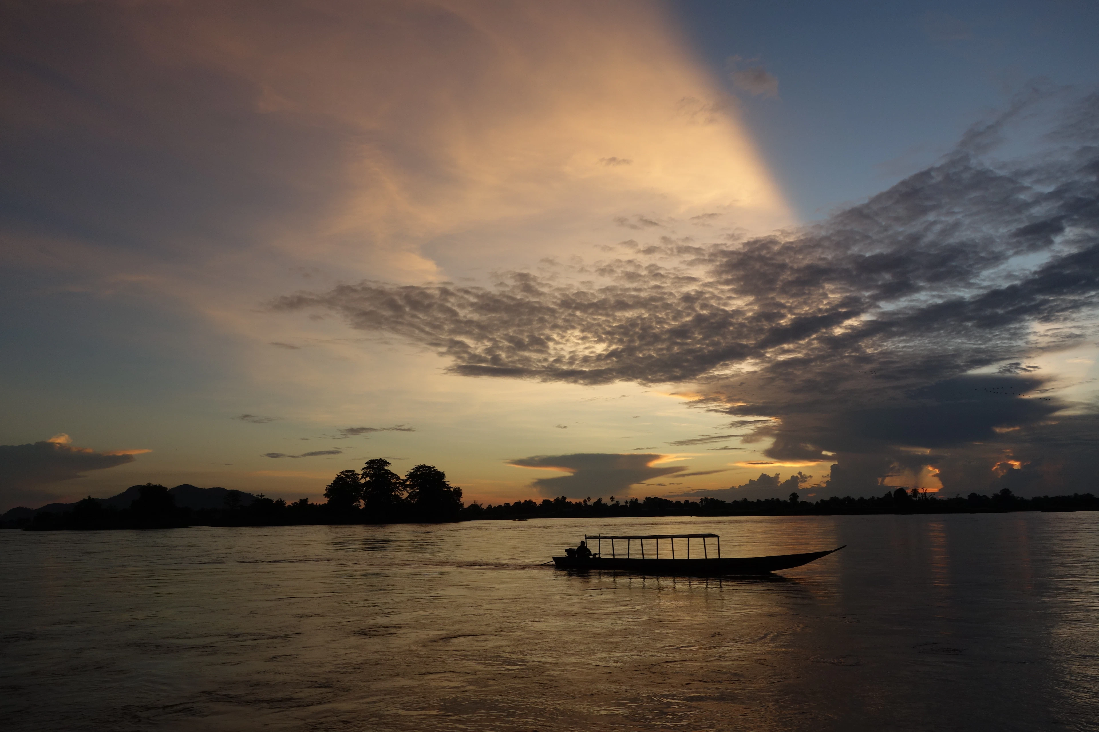 A wooden longtail boat silhouetted on a wide river at dusk beneath a shaft of golden light breaking through storm clouds.