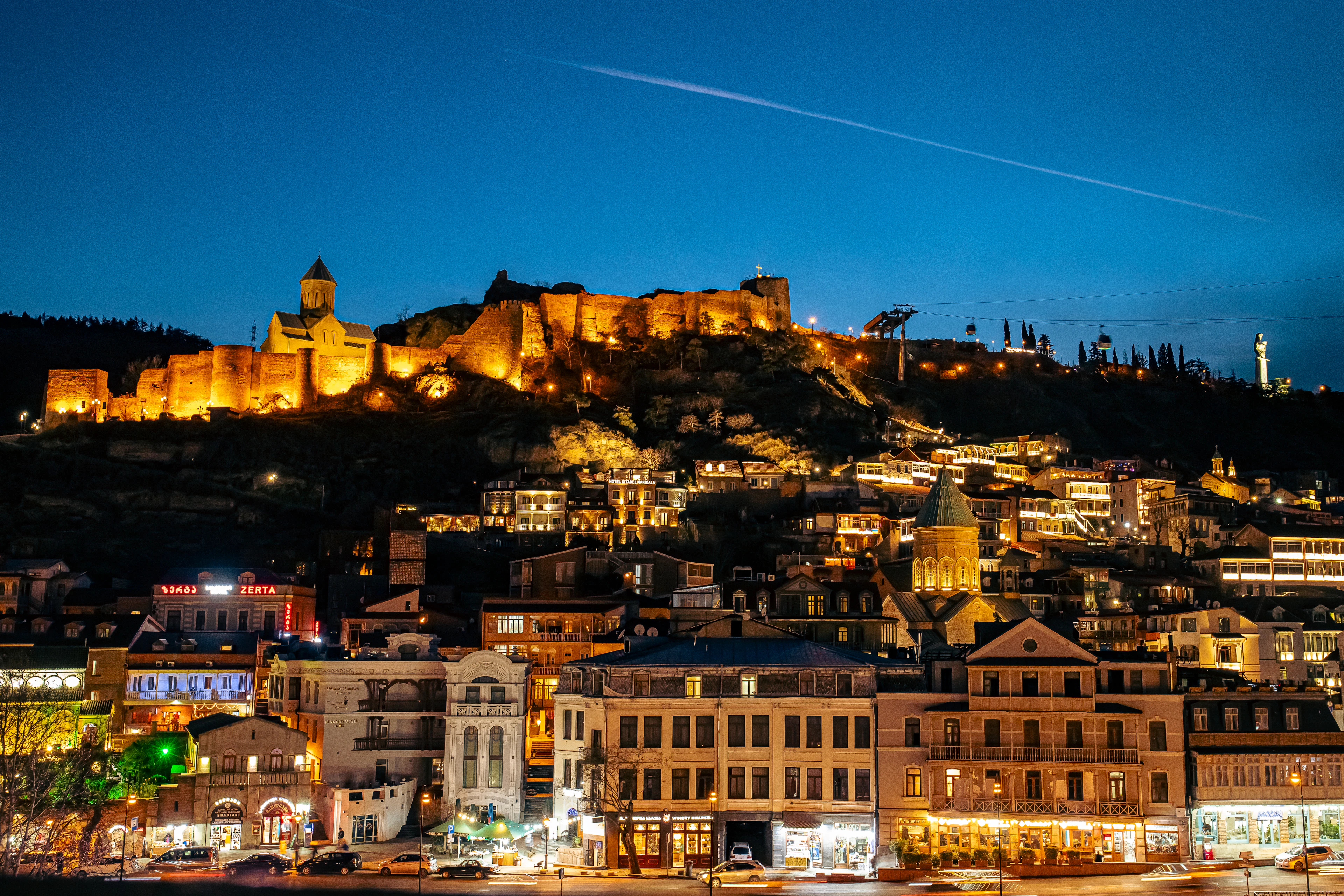 Narikala fortress floodlit along its ridge above Tbilisi at night, the lit houses, balconies and church dome of the old town spilling down the hillside beneath it under a deep blue sky.
