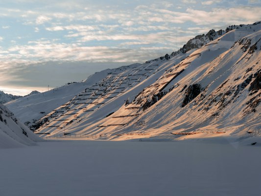Rows of avalanche barriers on a snow-covered slope above the valley, lit by low evening sun