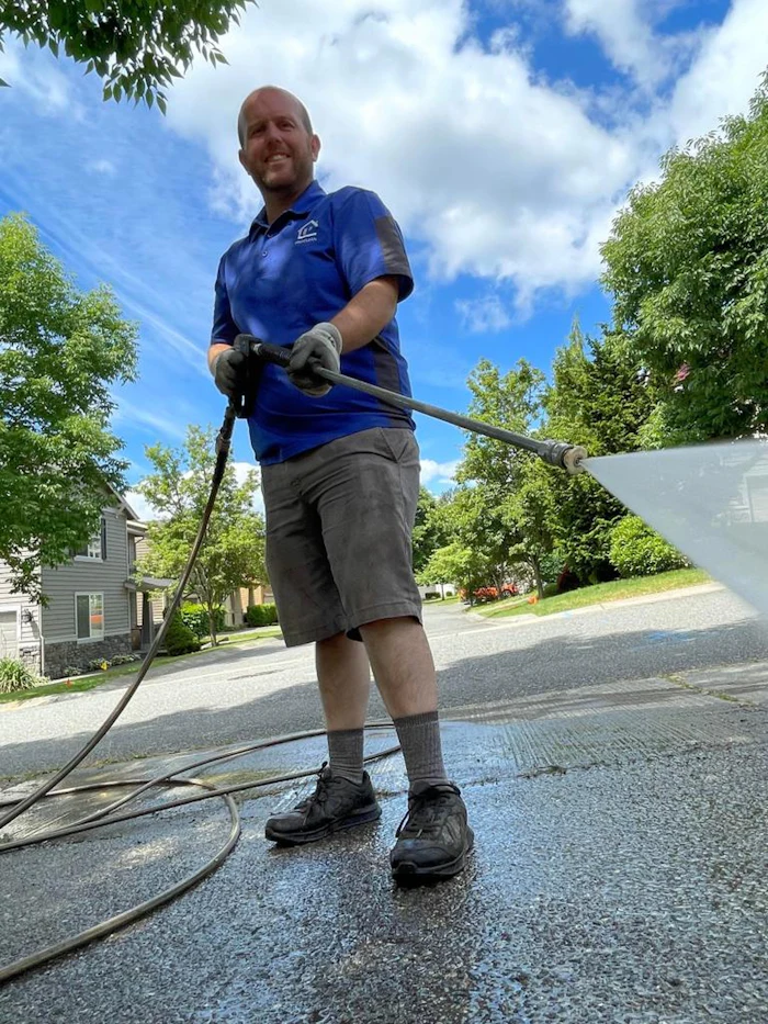 A technician in a blue shirt pressure washing a paved driveway.
