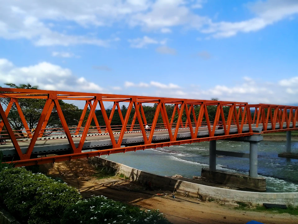 The image depicts a striking orange metal bridge spanning across a river, with a clear blue sky and fluffy white clouds above it. The bridge is supported by two sturdy concrete pillars on either side, creating an impressive architectural structure that stands out against the natural backdrop. On one side of the river, there are lush green trees providing a sense of tranquility to the scene. A few people can be seen walking along the bridge or standing near its base, adding a touch of human presence to this otherwise serene landscape.

The image is taken from an elevated perspective, offering a comprehensive view of the bridge and its surroundings. The colors in the photo are vibrant, with the orange of the bridge contrasting beautifully against the blue sky and green trees.