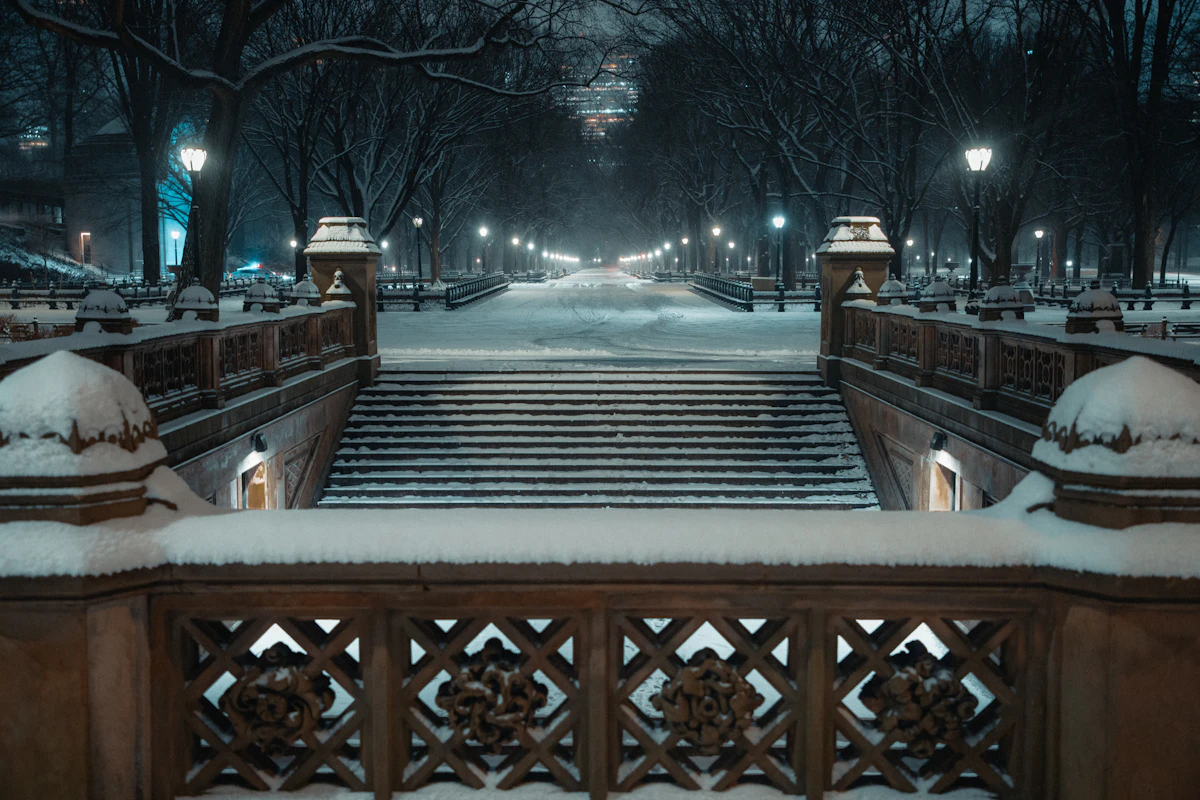 Snowy park path at night lined with benches and glowing lamps