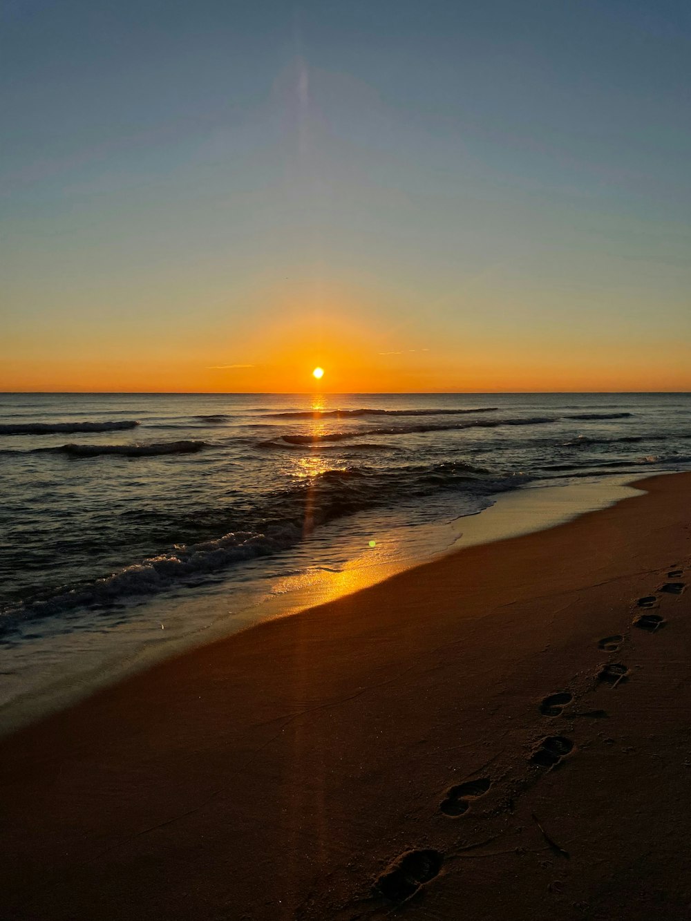 the sun is setting over the ocean with footprints in the sand