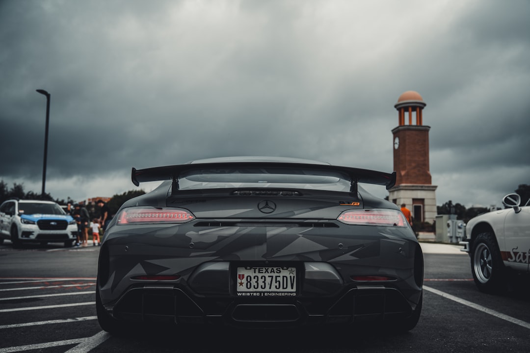 a black sports car parked in a parking lot, The image shows the rear view of a black Mercedes-AMG GT R parked in a lot. The car features a large rear wing and a camo-style wrap. The Texas license plate is visible, along with the car’s distinctive taillights and AMG badging. In the background, there are other vehicles, people, and a brick clock tower under a cloudy sky, indicating a car event or gathering.