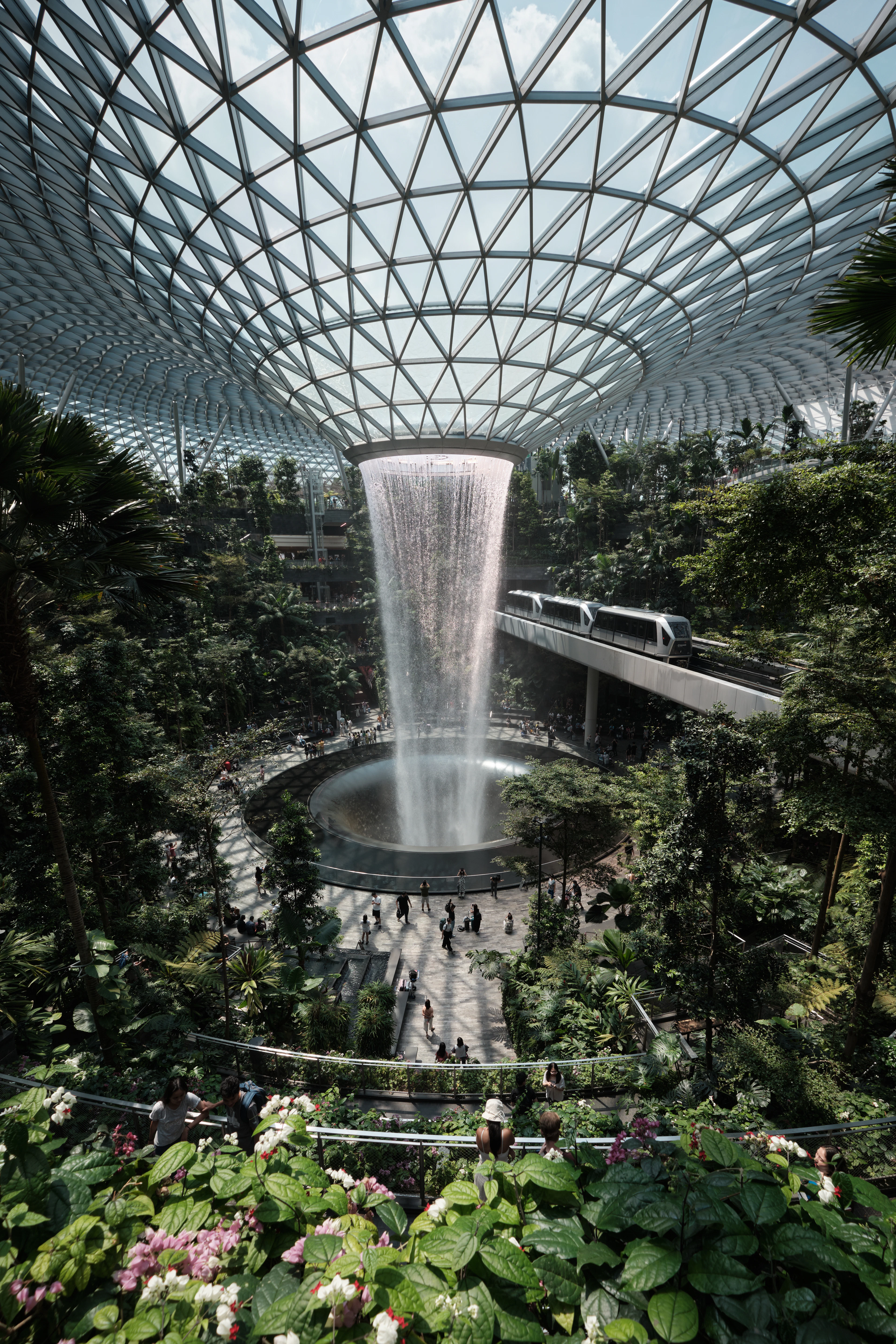 The Rain Vortex indoor waterfall pouring through the glass-and-steel dome of Jewel Changi Airport in Singapore, the Skytrain crossing the terraced indoor forest