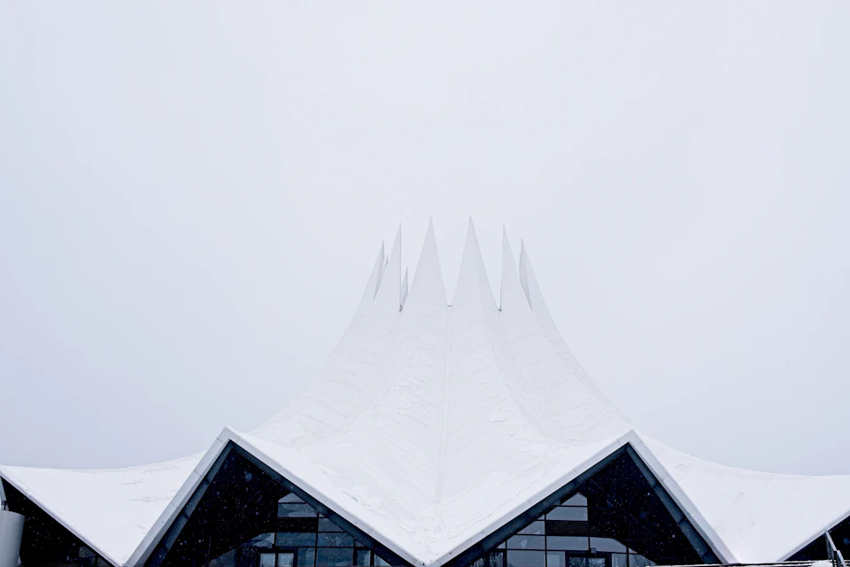 The image shows a large, white building with a pointed roof and multiple spires on its sides. The building is situated against an overcast sky, which casts a soft light that highlights the architectural details. The perspective of the photo is from below, looking up at the imposing structure.