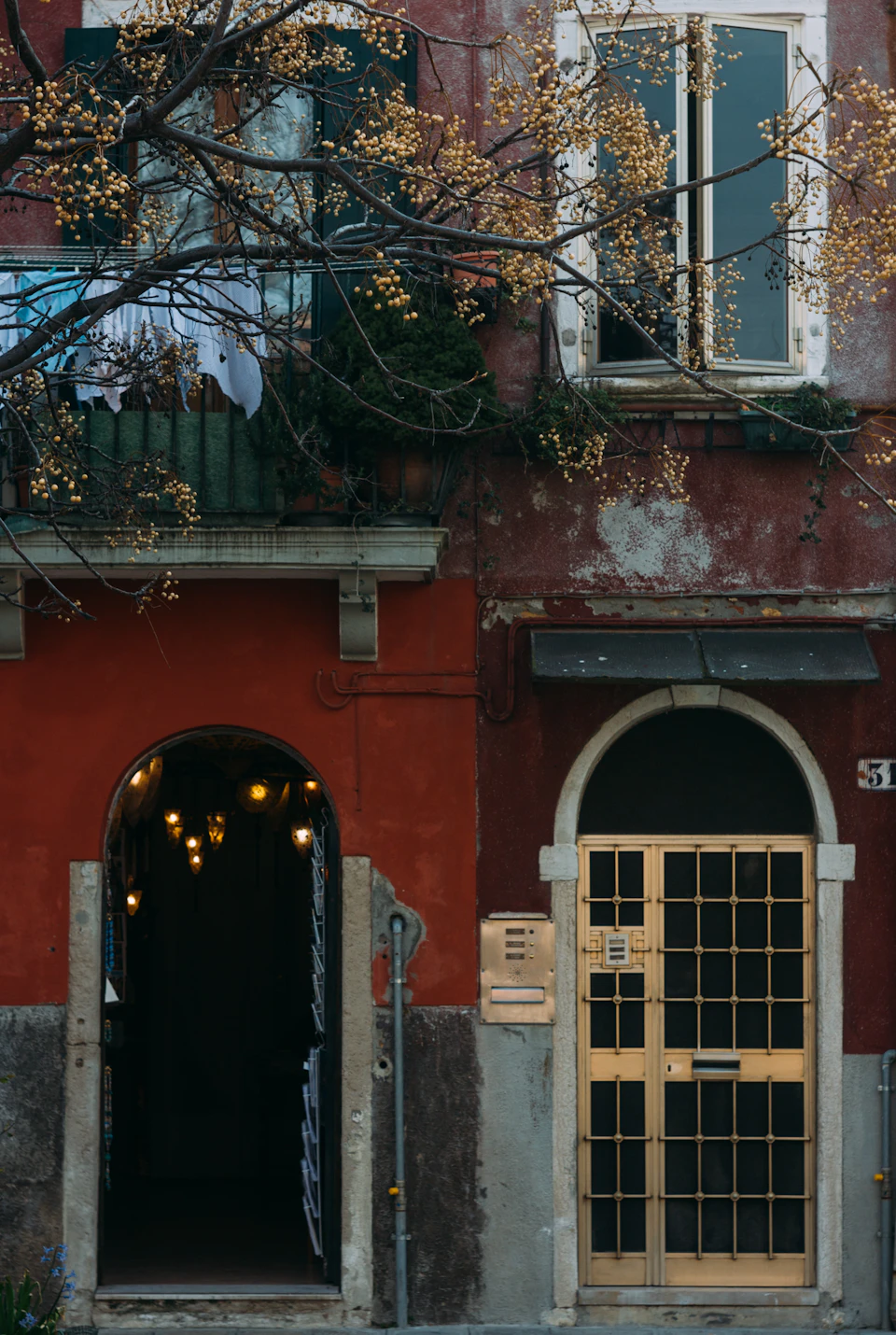 The image depicts a charming red building with an arched doorway and a balcony on the second floor, both adorned with hanging lights that cast a warm glow onto the scene below. The building's facade is painted in a rich shade of red, contrasting beautifully with the clear blue sky visible through the windows. The perspective of the photo is from street level, looking up at the building, which adds to its grandeur and prominence.

In front of the building, there are two potted plants - one on each side of the entrance, adding a touch of greenery to the scene. A sign can be seen hanging above the door, though its contents are not discernible from this angle.