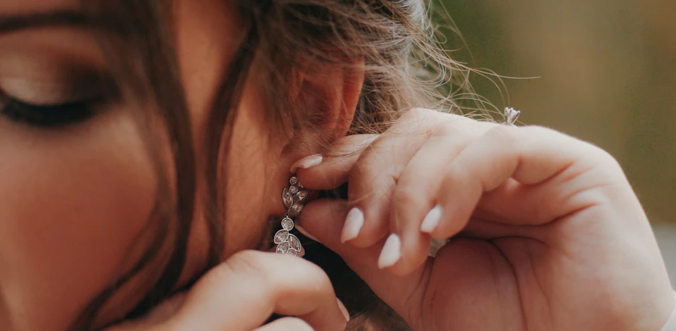 The image shows a close-up view of a woman's ear, with her left hand gently holding a pair of diamond earrings. The earrings are large and shiny, reflecting light off their surfaces. The woman is wearing white nail polish on her fingers, adding to the overall elegance of the scene.