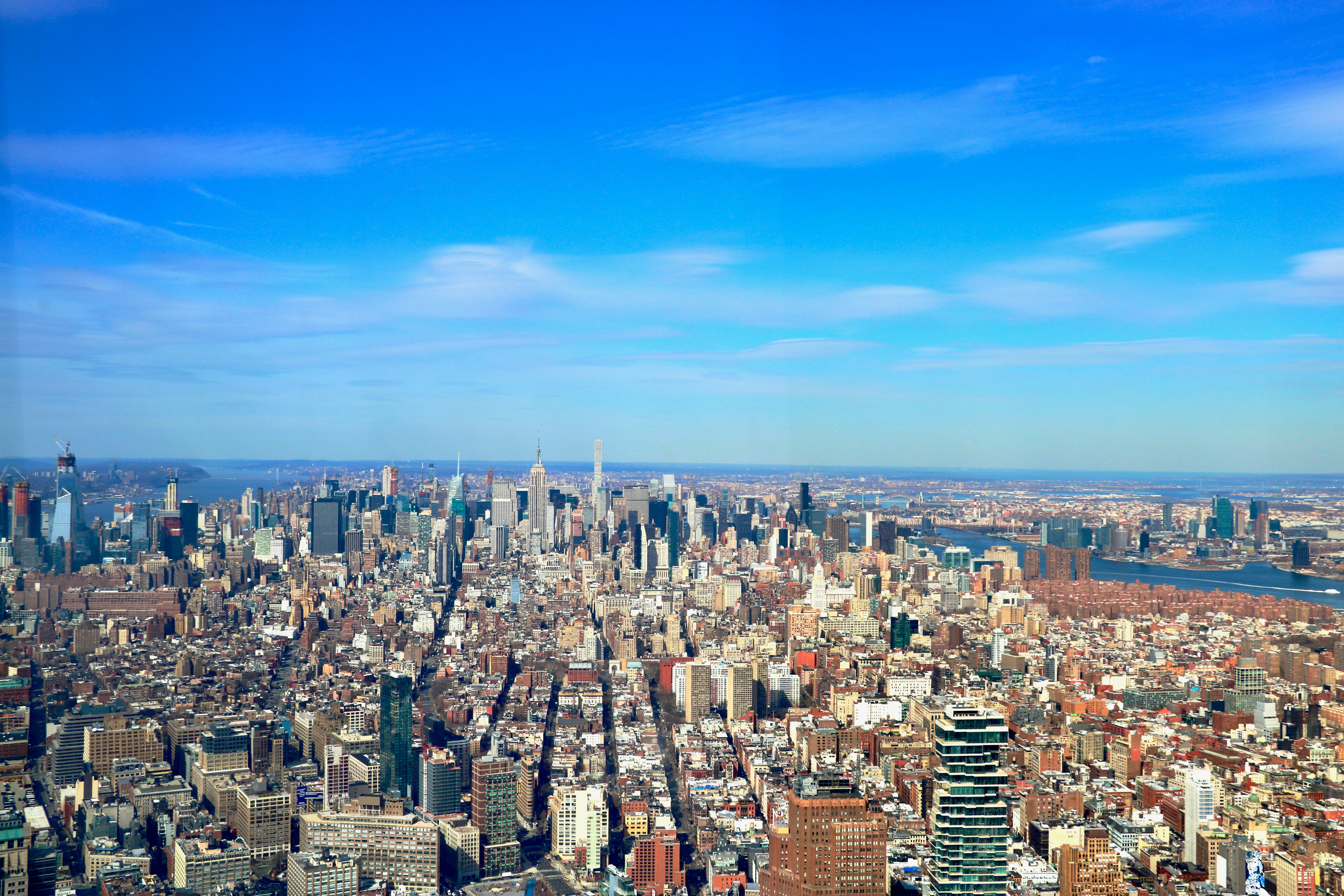 an aerial view of a city with tall buildings, Pictured here is a panoramic aerial view of Manhattan from the One World Observatory in Lower Manhattan. Some neighborhoods that can be seen here are the East Village, the Lower East Side, Tribeca, and Union Square with the Empire State building in midtown and the east river in the far distance.