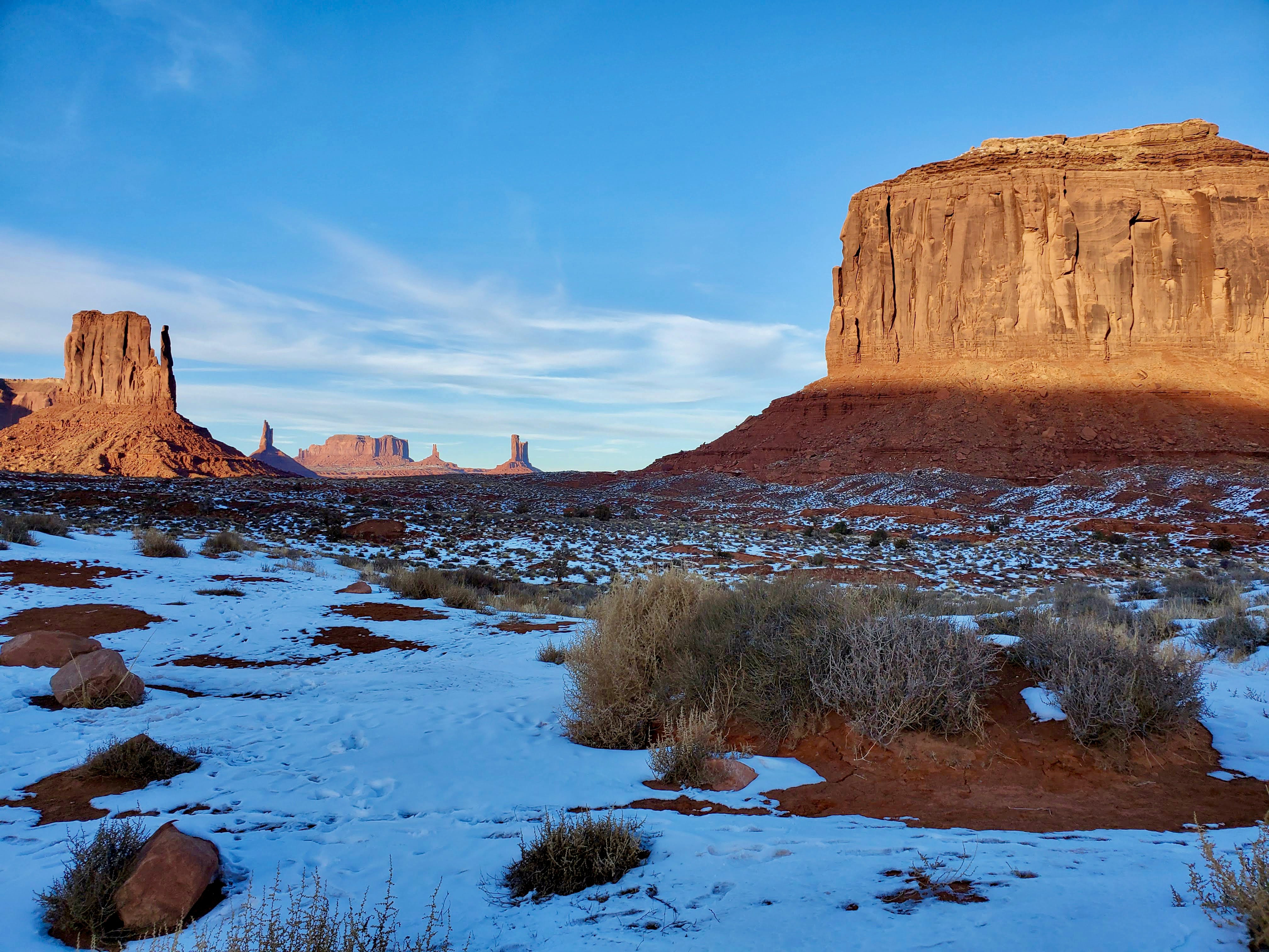 a snow covered field with a mountain in the background, Sunset at Monument Valley. situated on the Arizona-Utah border in the United States, Monument Valley is an iconic and otherworldly landscape characterized by its towering sandstone buttes, mesas, and dramatic desert vistas. This extraordinary geological formation has become synonymous with the American West and has served as the backdrop for countless Western films and photographs, evoking a sense of rugged beauty and timelessness.